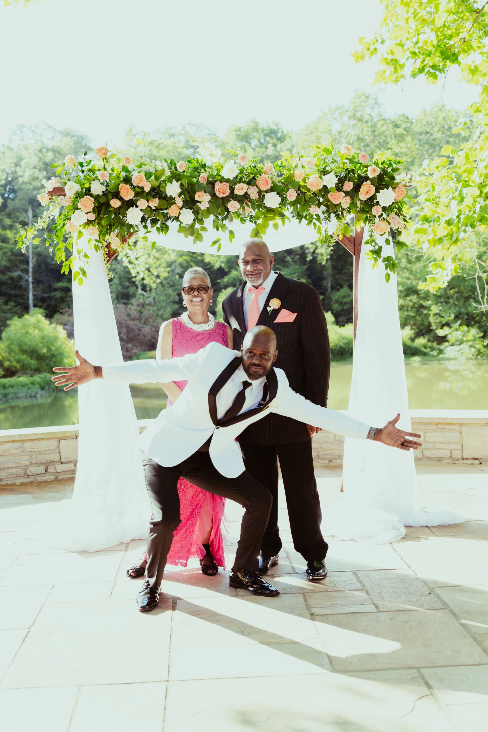 A man in a tuxedo is posing for a picture with his arms outstretched in front of a floral arch.