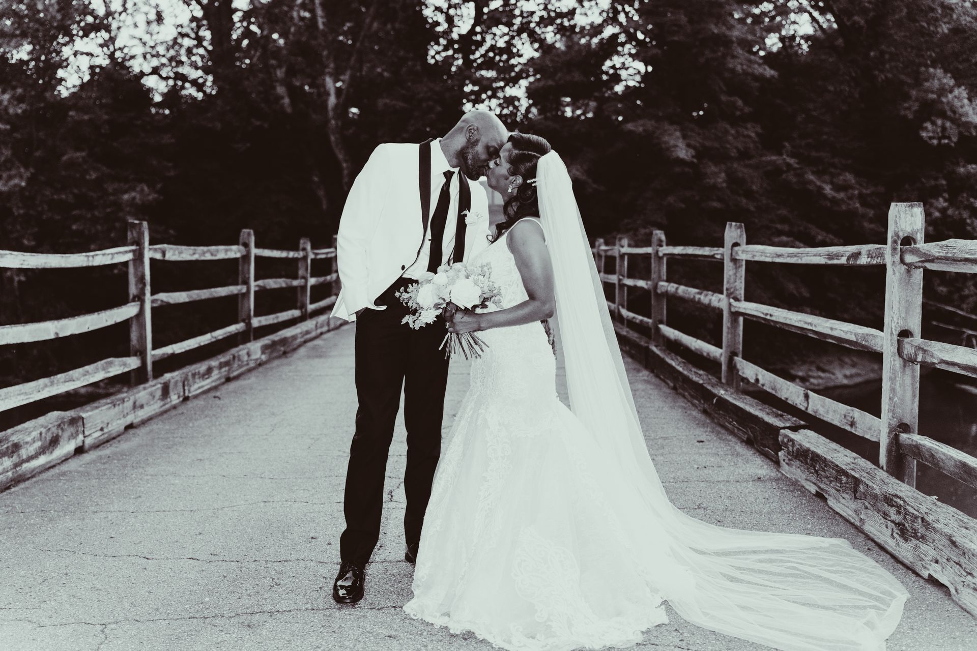 A black and white photo of a bride and groom kissing on a bridge