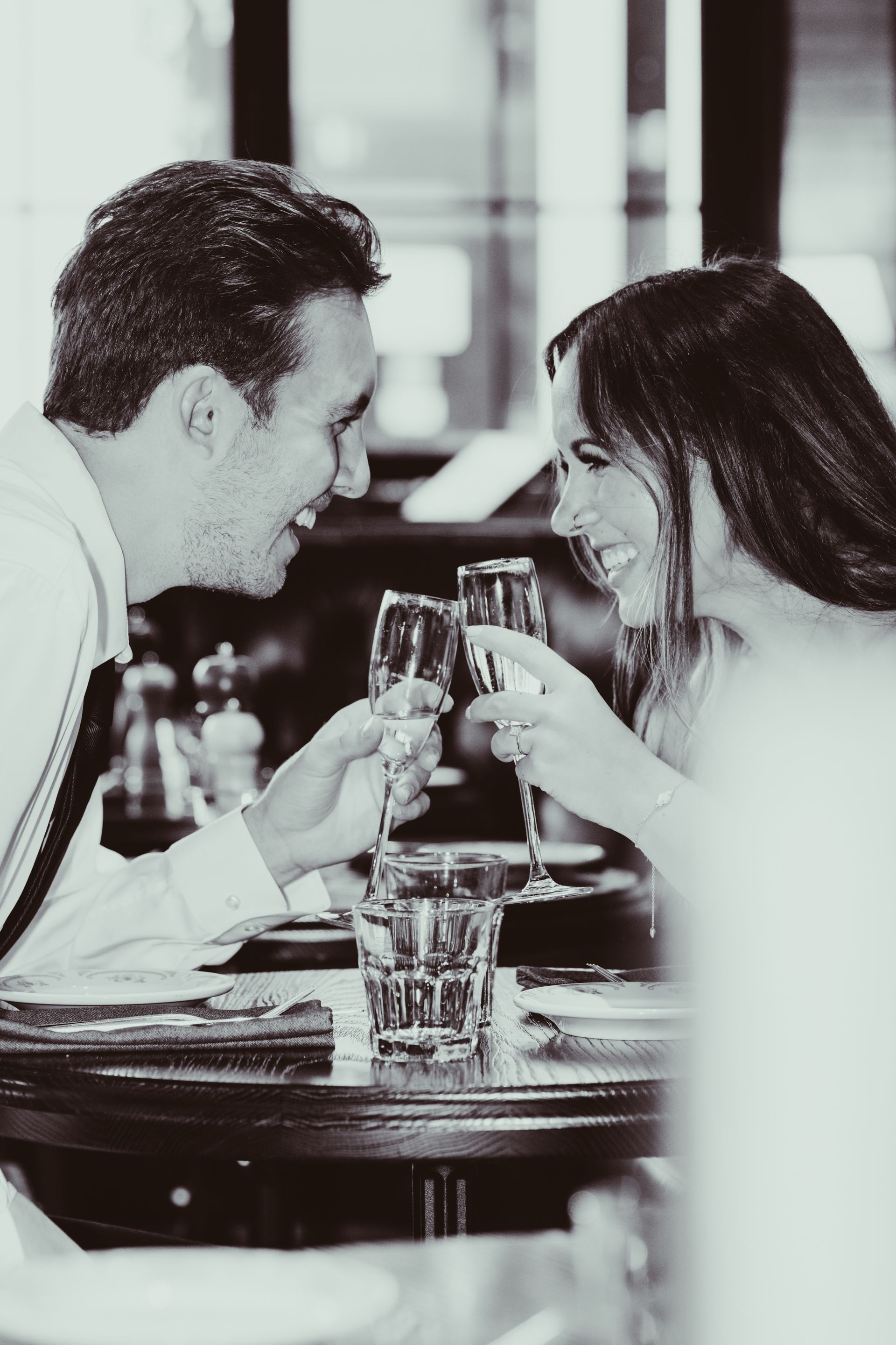 Couple toasting champagne at restaurant table, smiling, black and white.