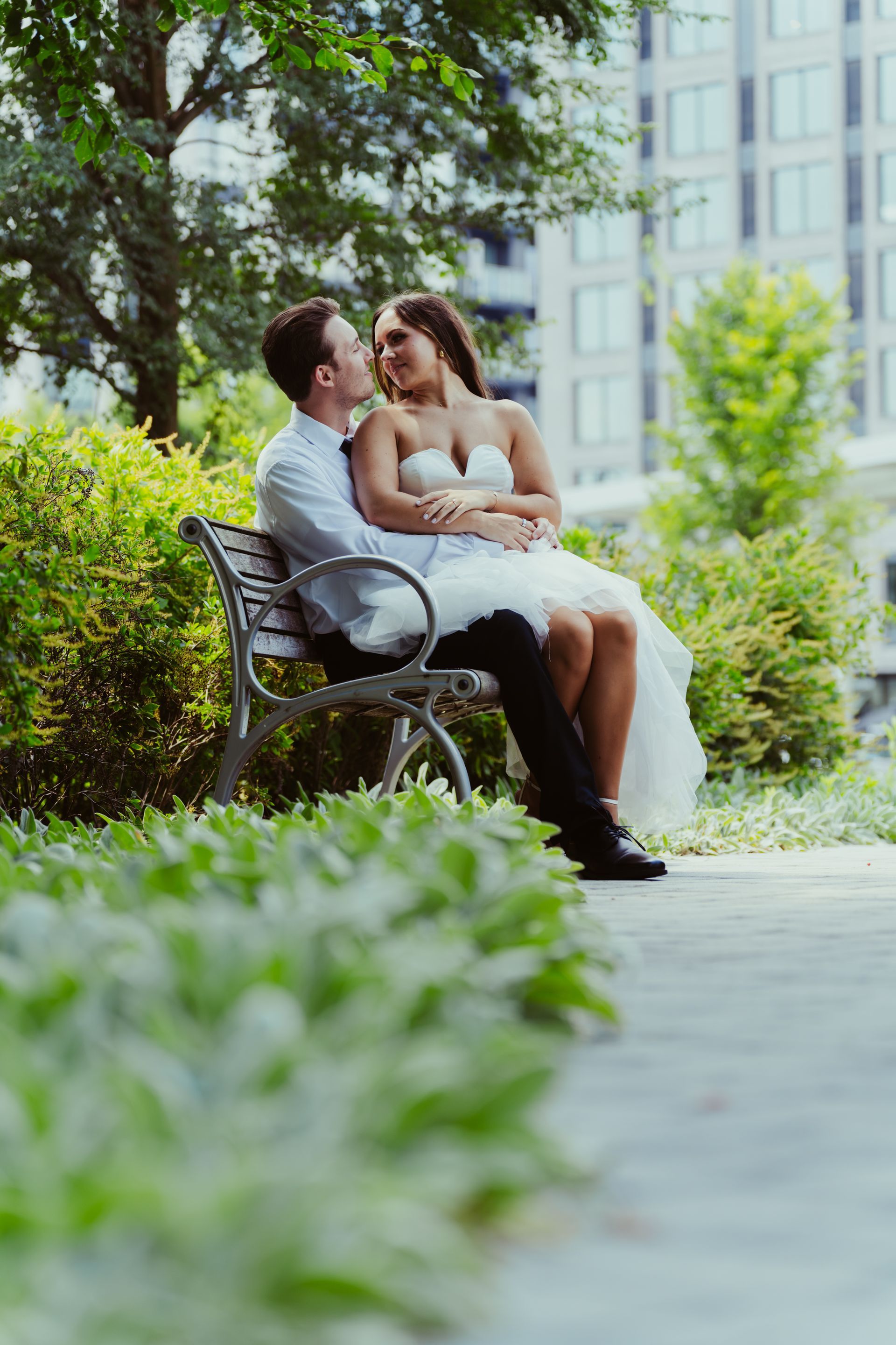 Couple in wedding attire on a park bench, looking at each other, with greenery and a city building in the background.
