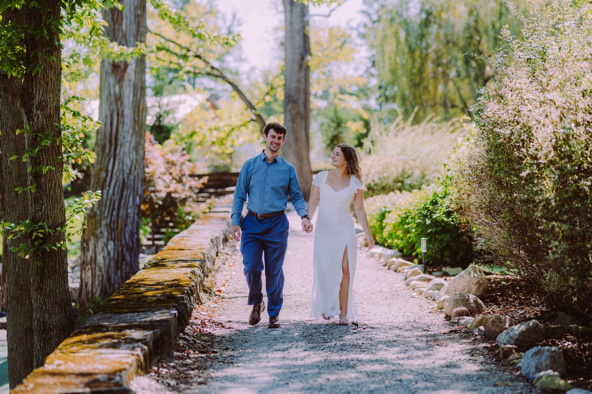 Couple walking hand-in-hand on a stone path, smiles, sunny day, surrounded by trees and greenery.