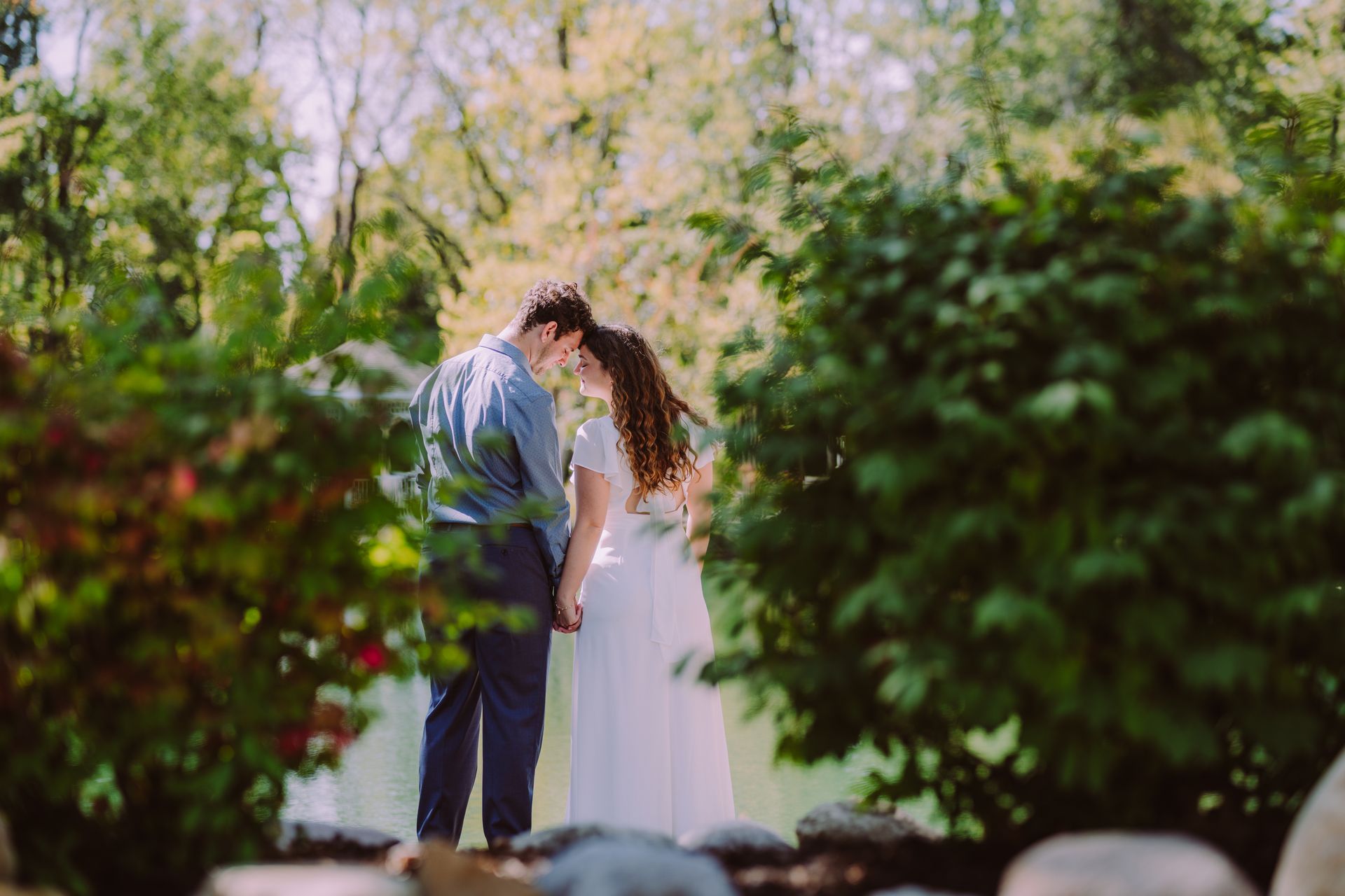 Couple embracing in a garden, holding hands, with a lake and foliage in the background.