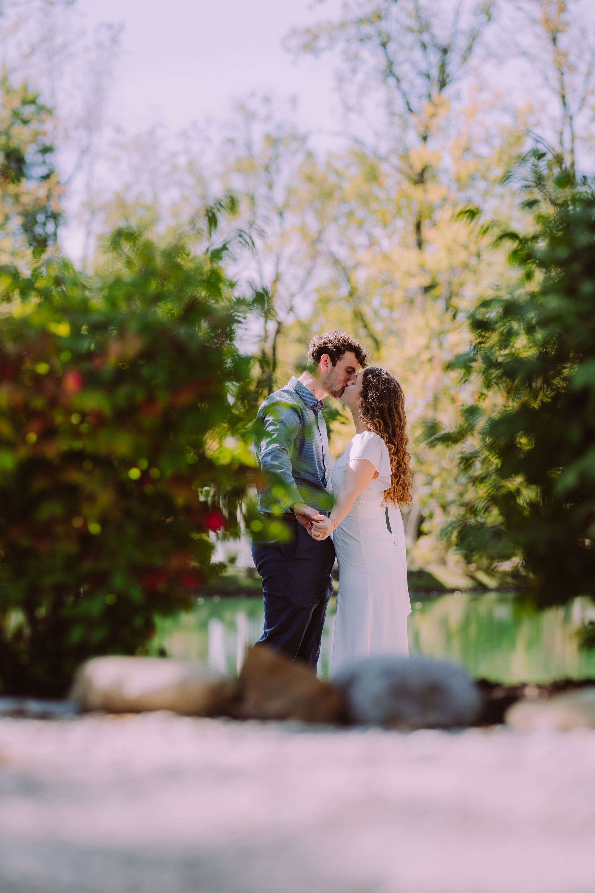 Couple in embrace, holding hands, about to kiss. Standing near trees and pond on sunny day.
