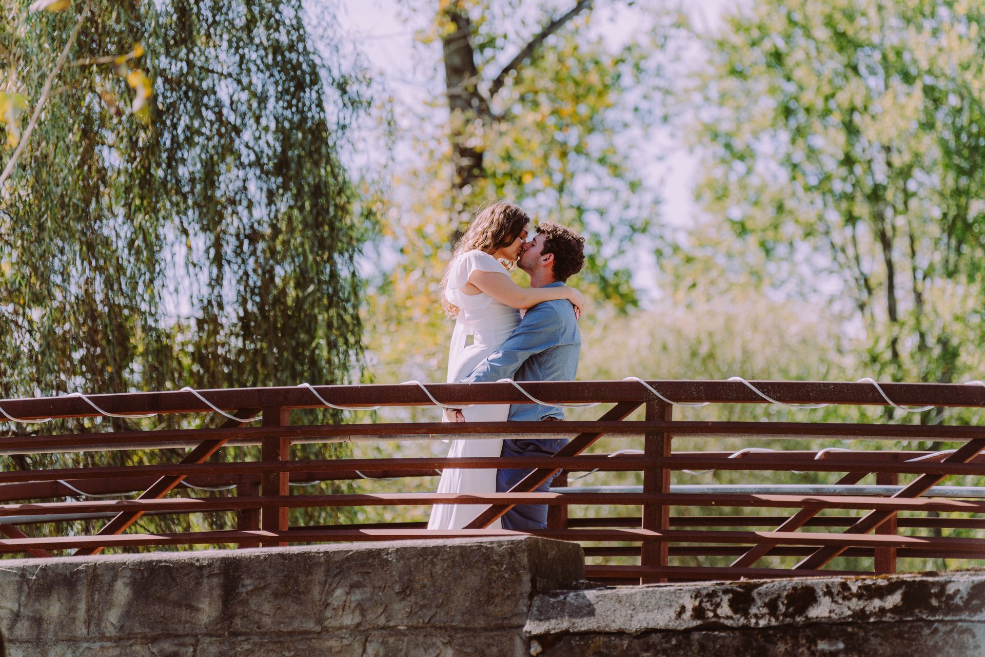 Couple embraces on a bridge, kissing. Wooden railing, trees with yellow leaves in background.