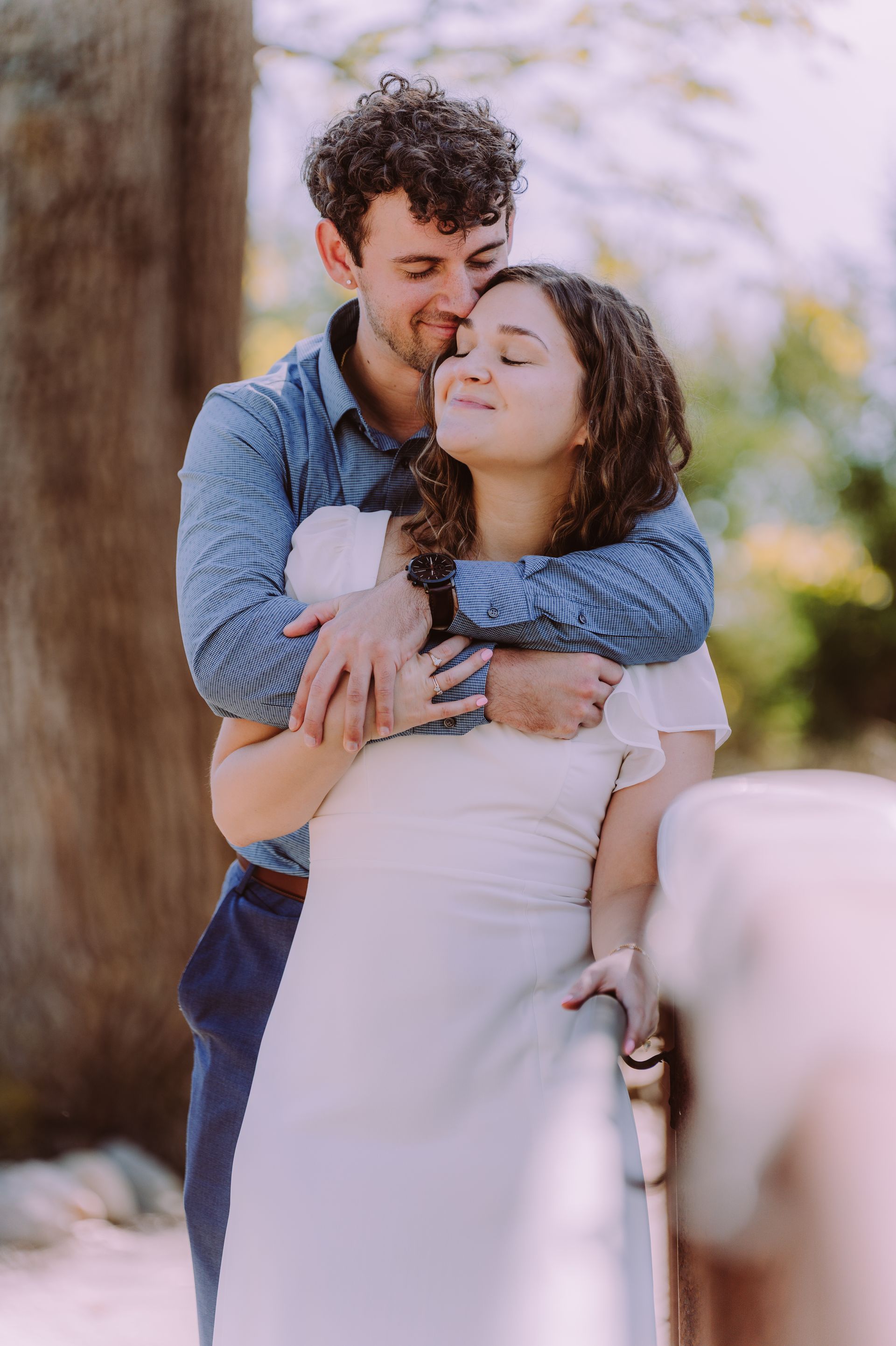 Man hugging woman, both smiling with eyes closed; outdoors, near a wooden railing.