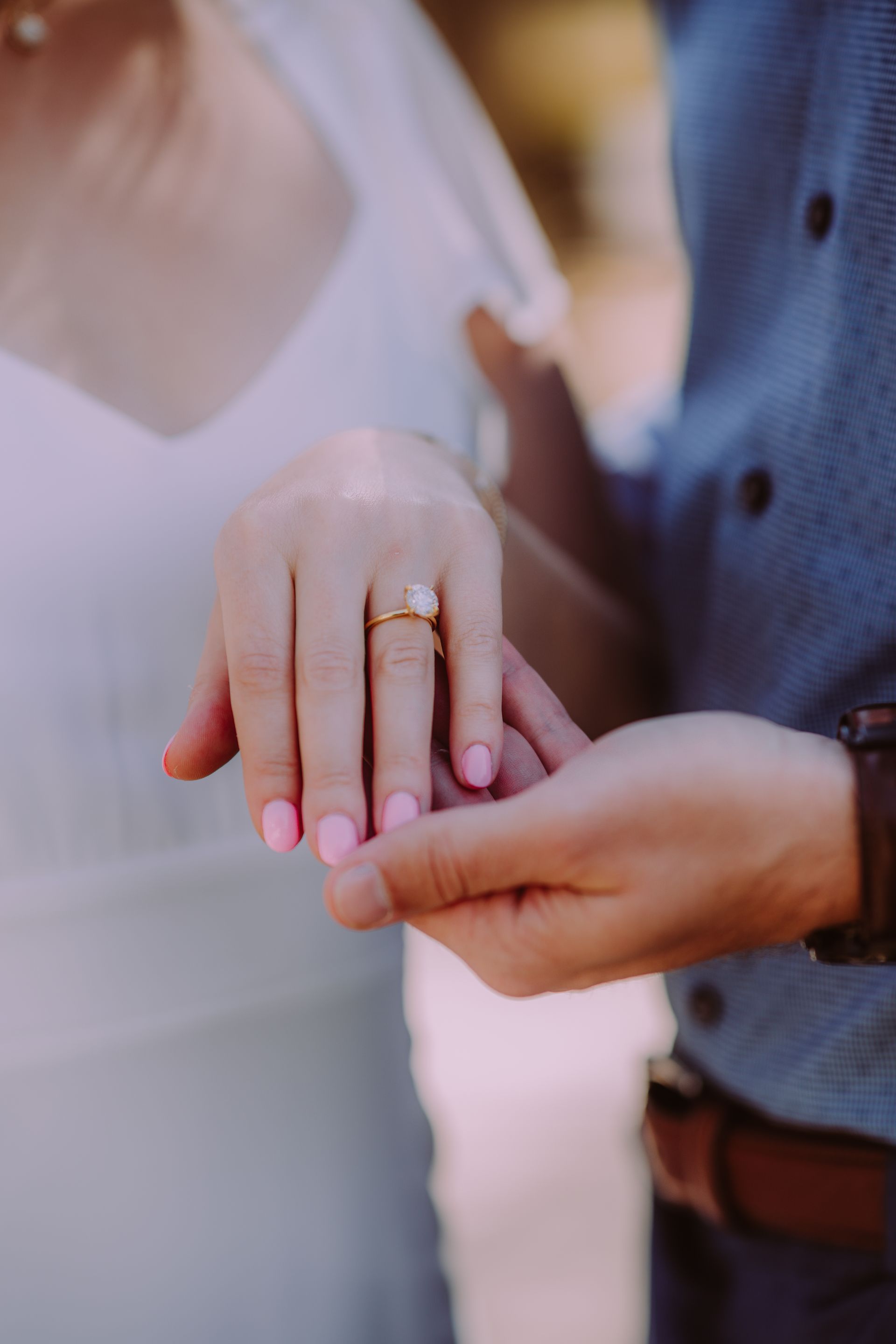 Person's hand with engagement ring held by another person's hand, blue shirt, pink nail polish.