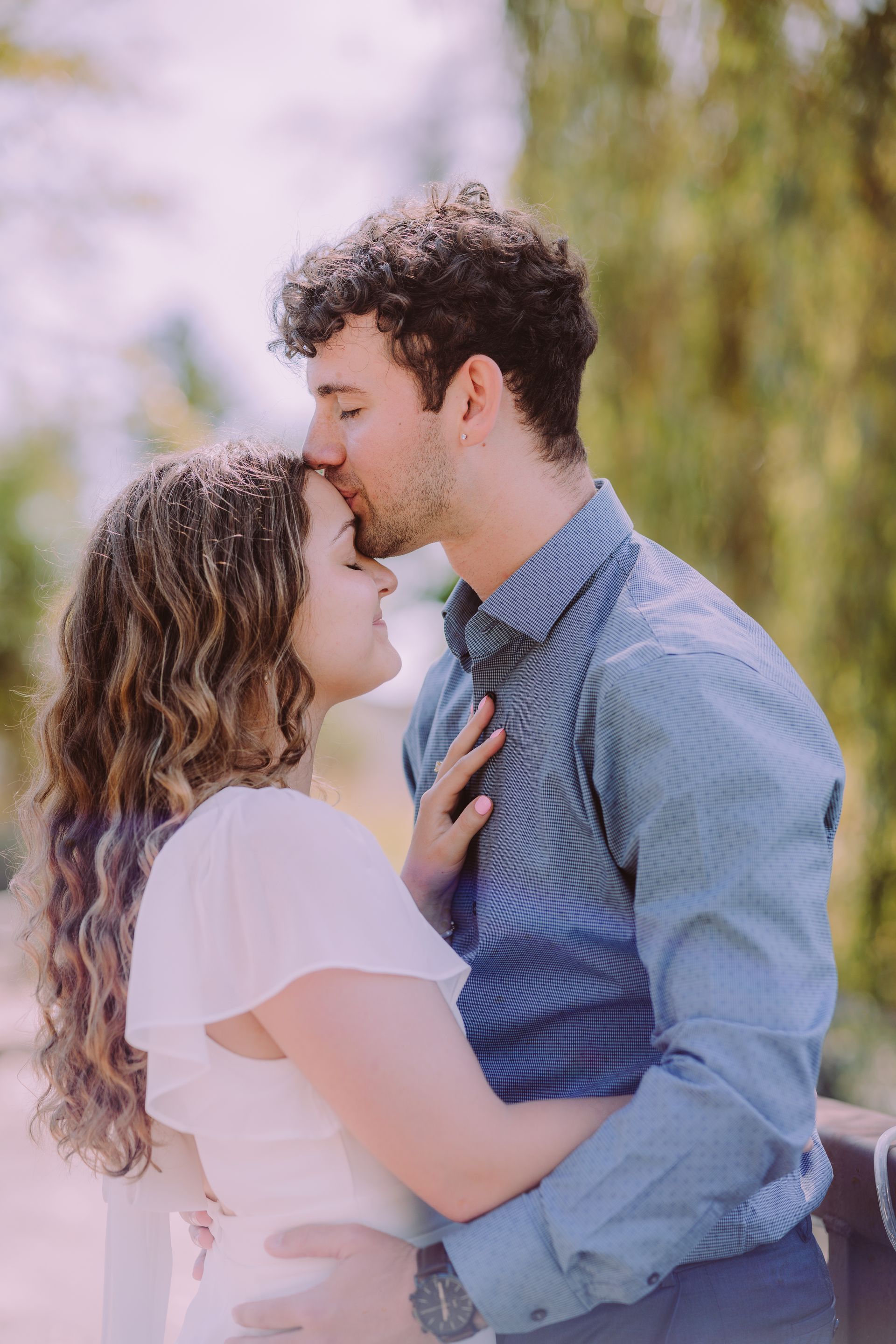 Man kisses woman's forehead; they embrace outdoors near greenery.