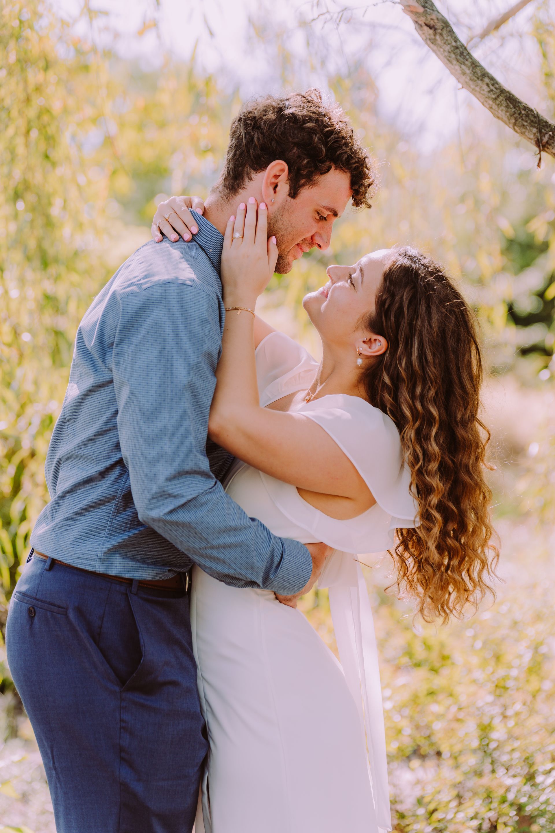 Couple embraces outdoors; woman touches man's face, both smiling. Soft sunlight, foliage background.