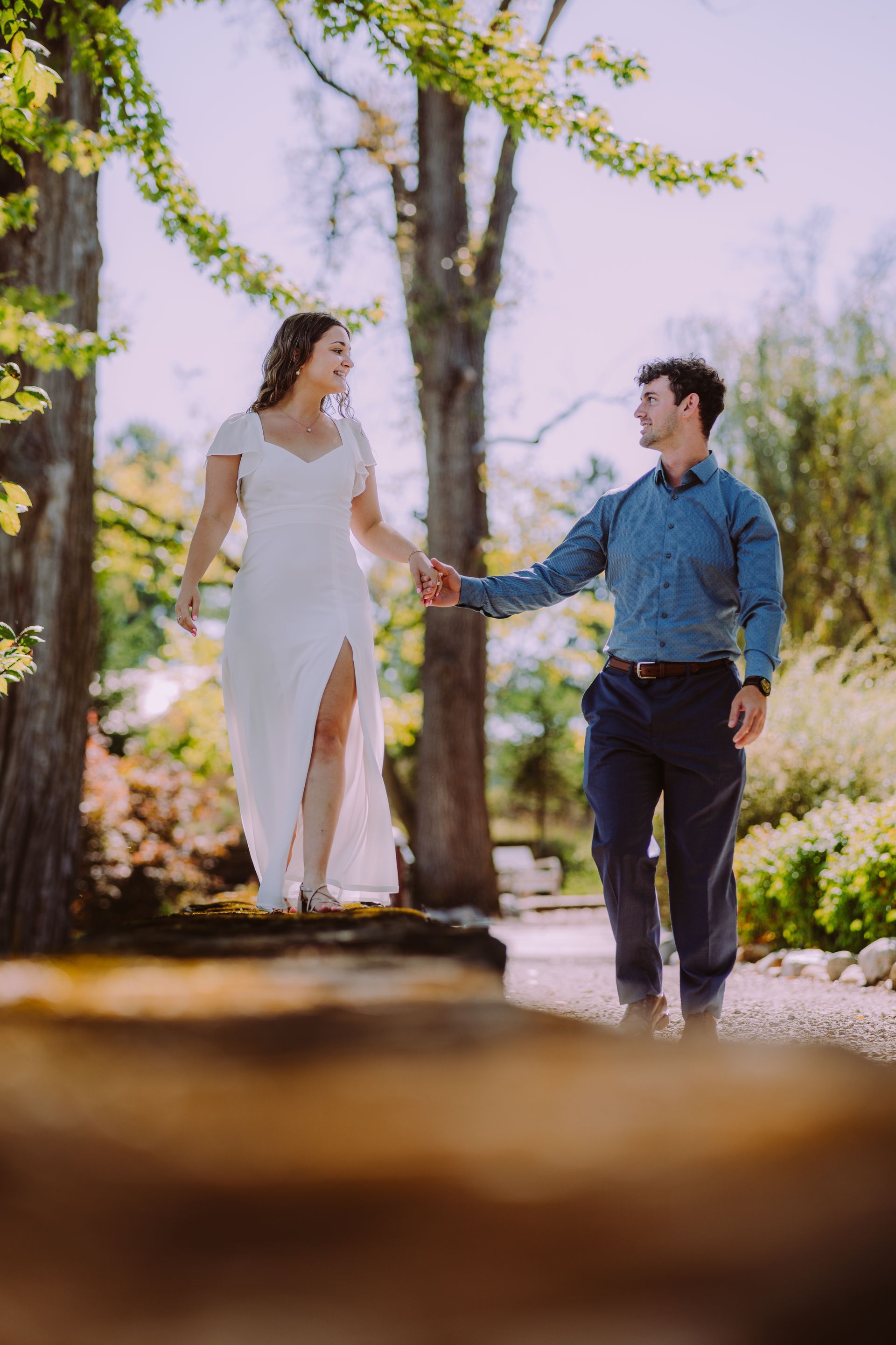 Couple holds hands, smiling, in a sunlit outdoor garden setting. Woman wears white dress, man a blue shirt and navy pants.