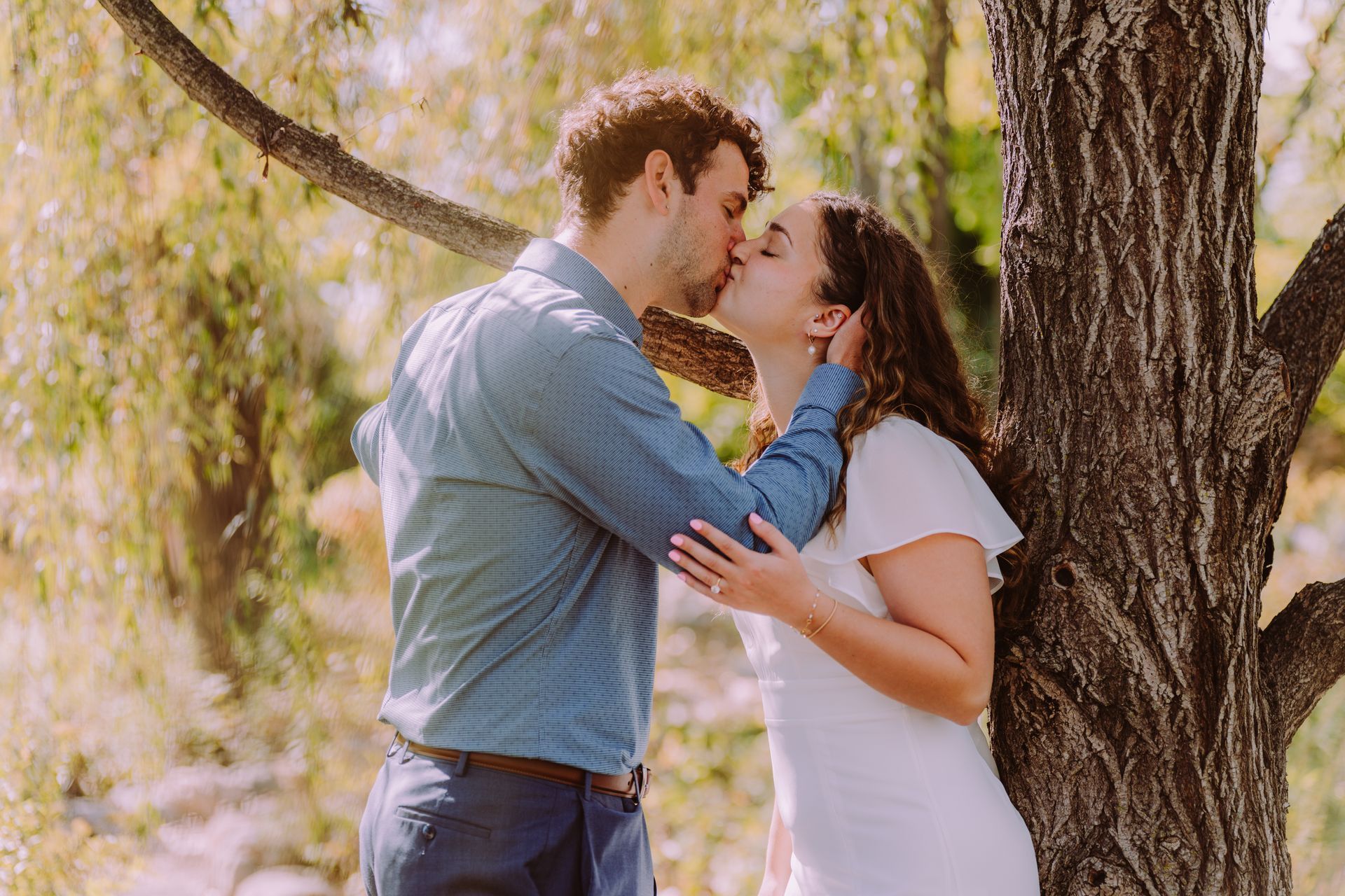 Couple kissing near a tree, outdoors in sunlight. The woman wears a white dress and the man a blue shirt.