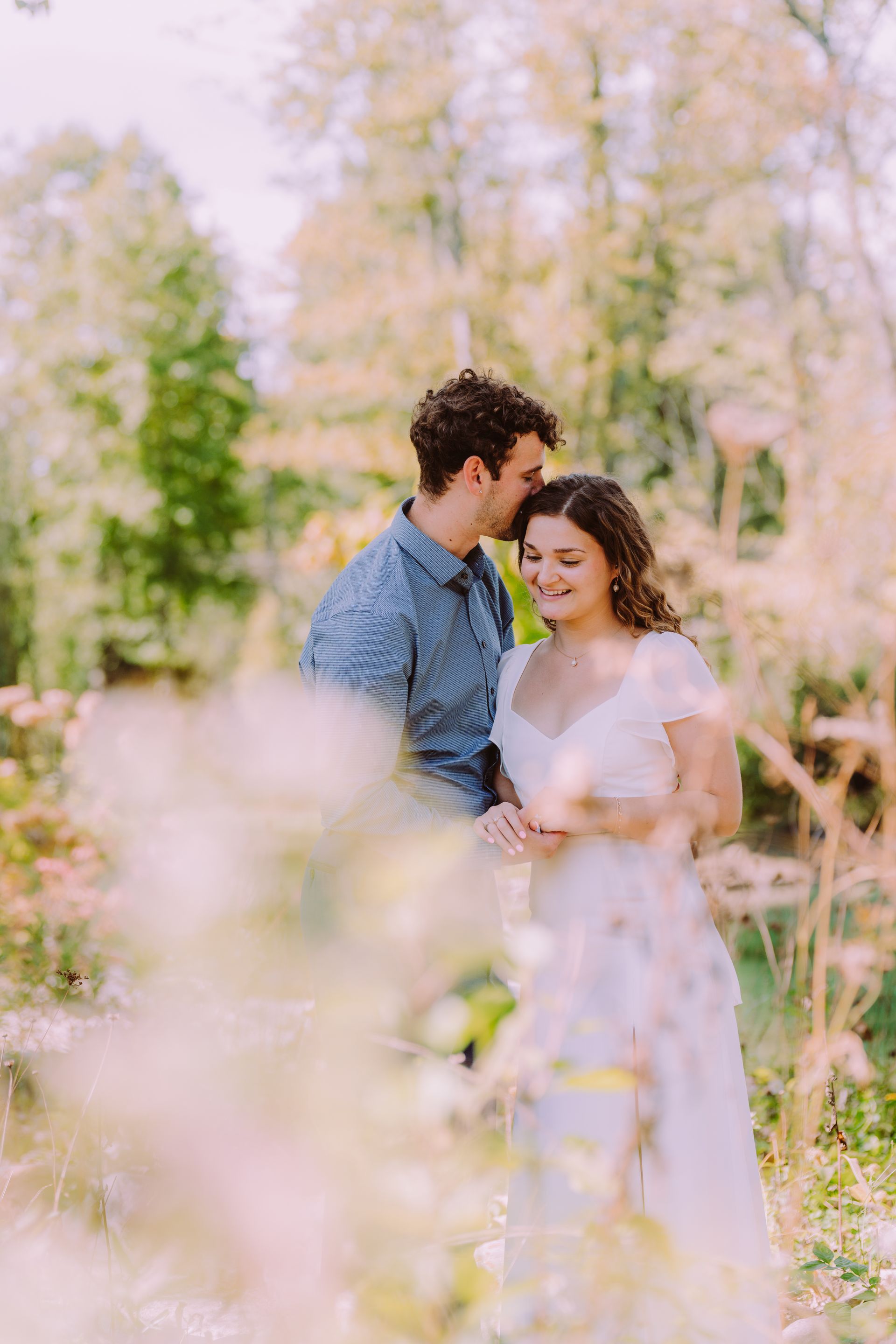 Couple embracing in a sunlit garden; man kisses woman's forehead. Soft focus, pastel colors, natural light.