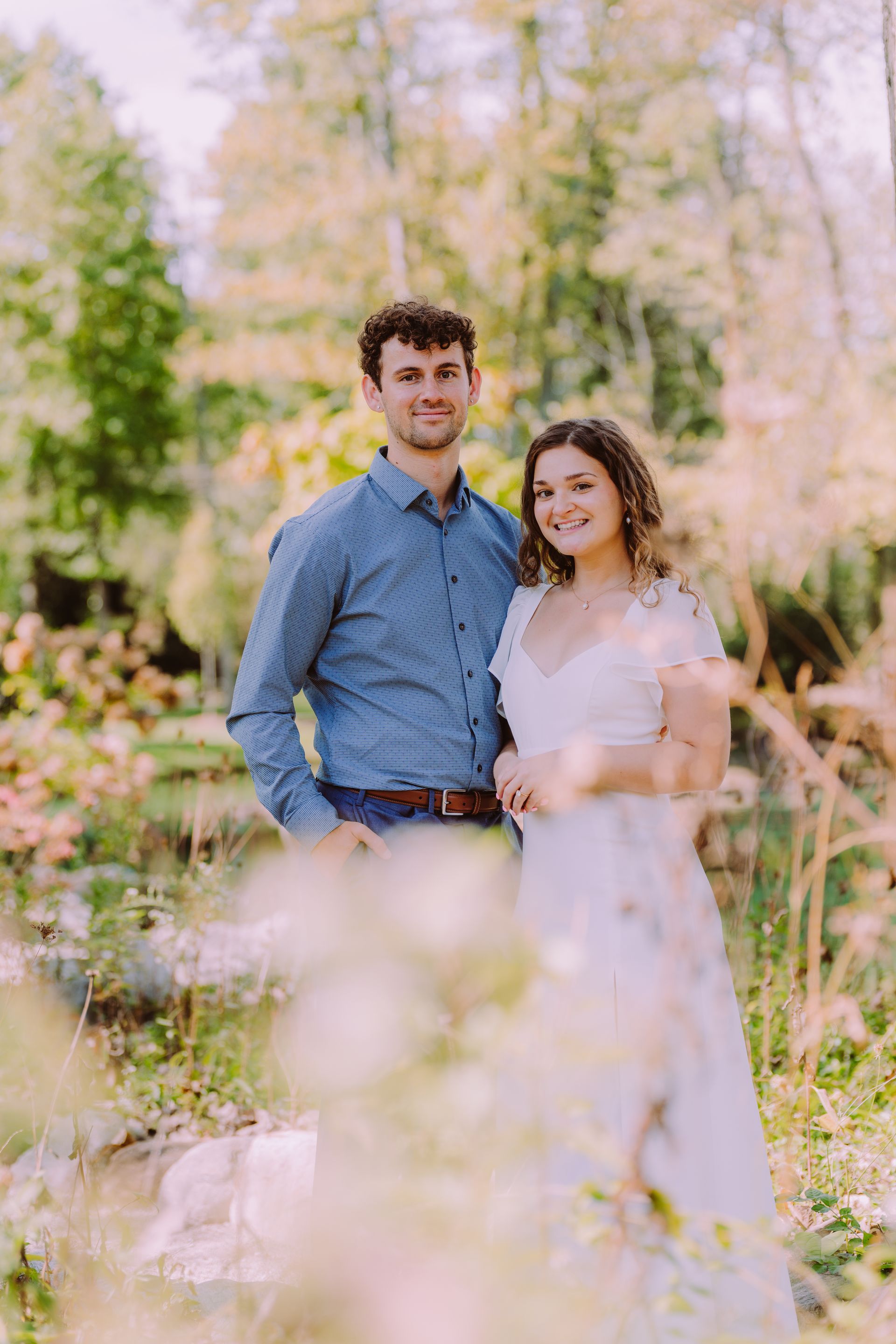 Couple standing outdoors, posing for a photo. Man in blue shirt, woman in white dress. Foliage in background.