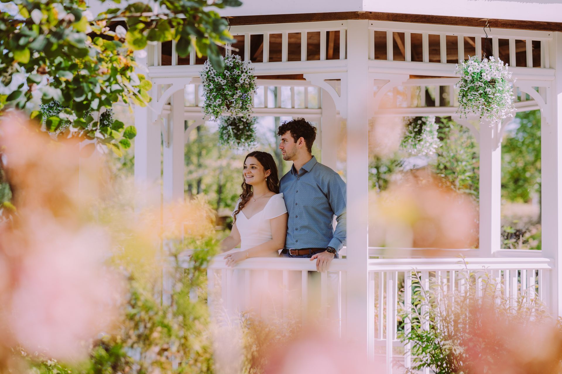 Couple in white gazebo, embracing, looking off into the distance, surrounded by blurred flowers and greenery.