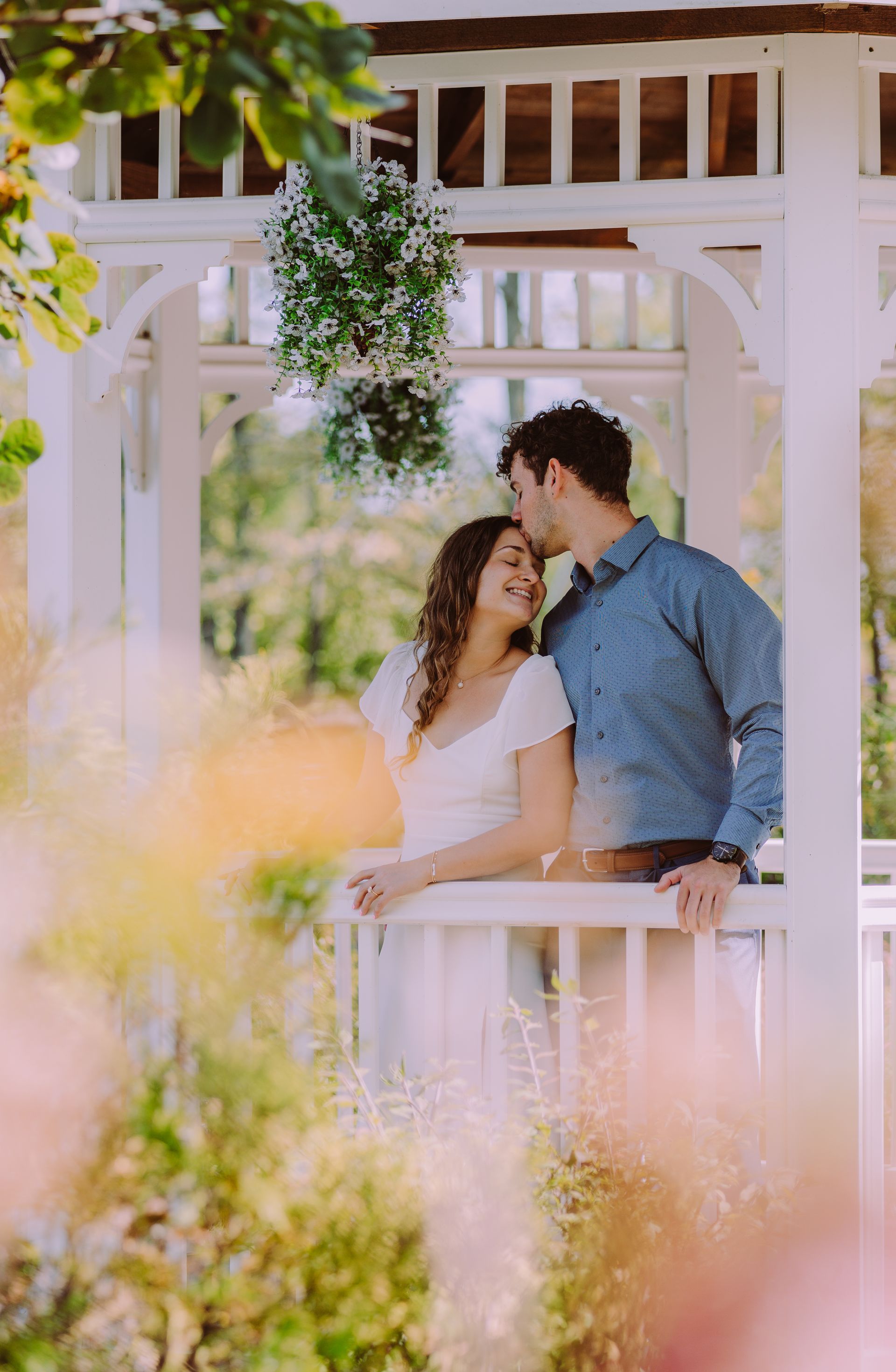 Couple in white gazebo, man kissing woman's forehead, surrounded by greenery.