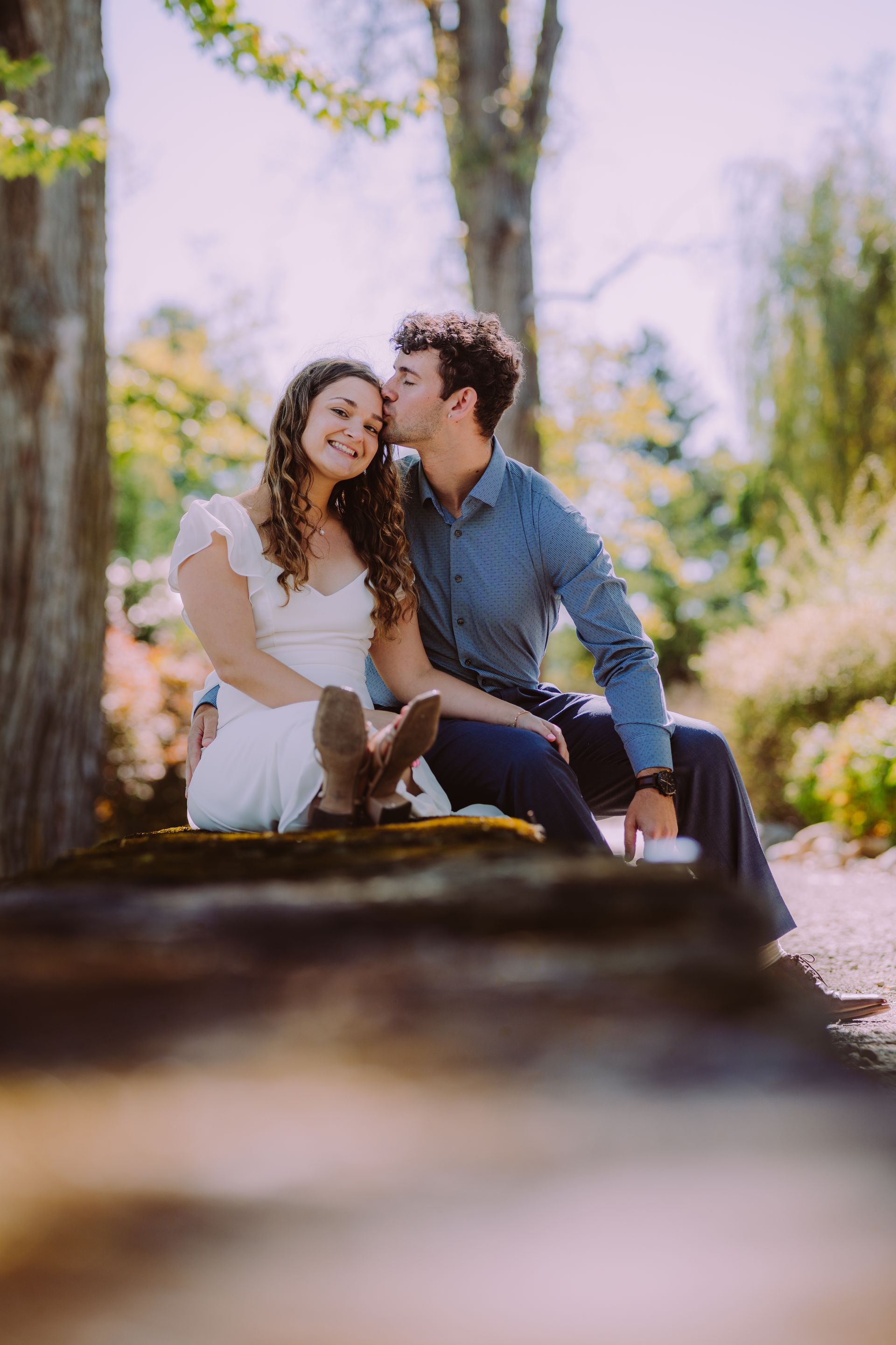 Couple seated outdoors, the man kissing the woman's forehead. They are smiling in a sunny, garden setting.
