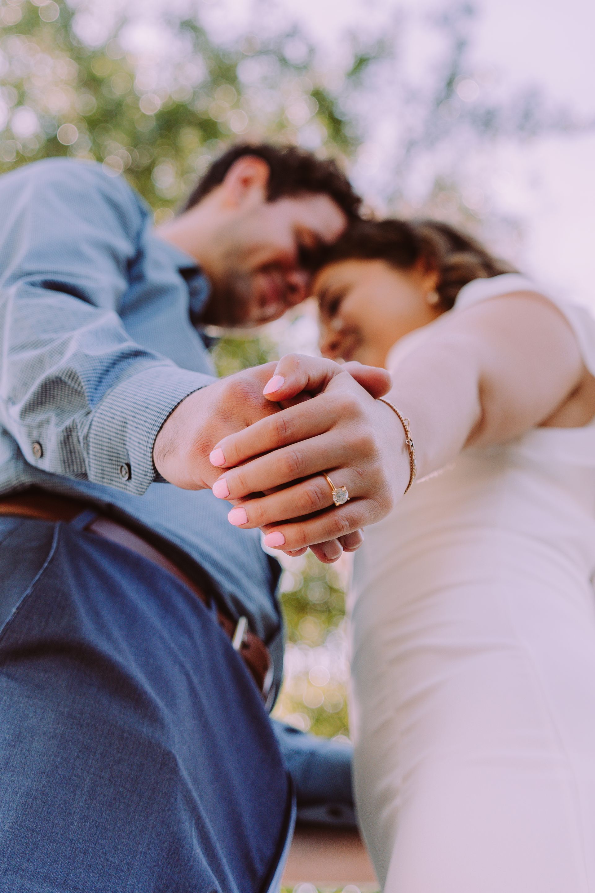 Couple holding hands, heads touching, celebrating. Engagement ring visible, outdoors.