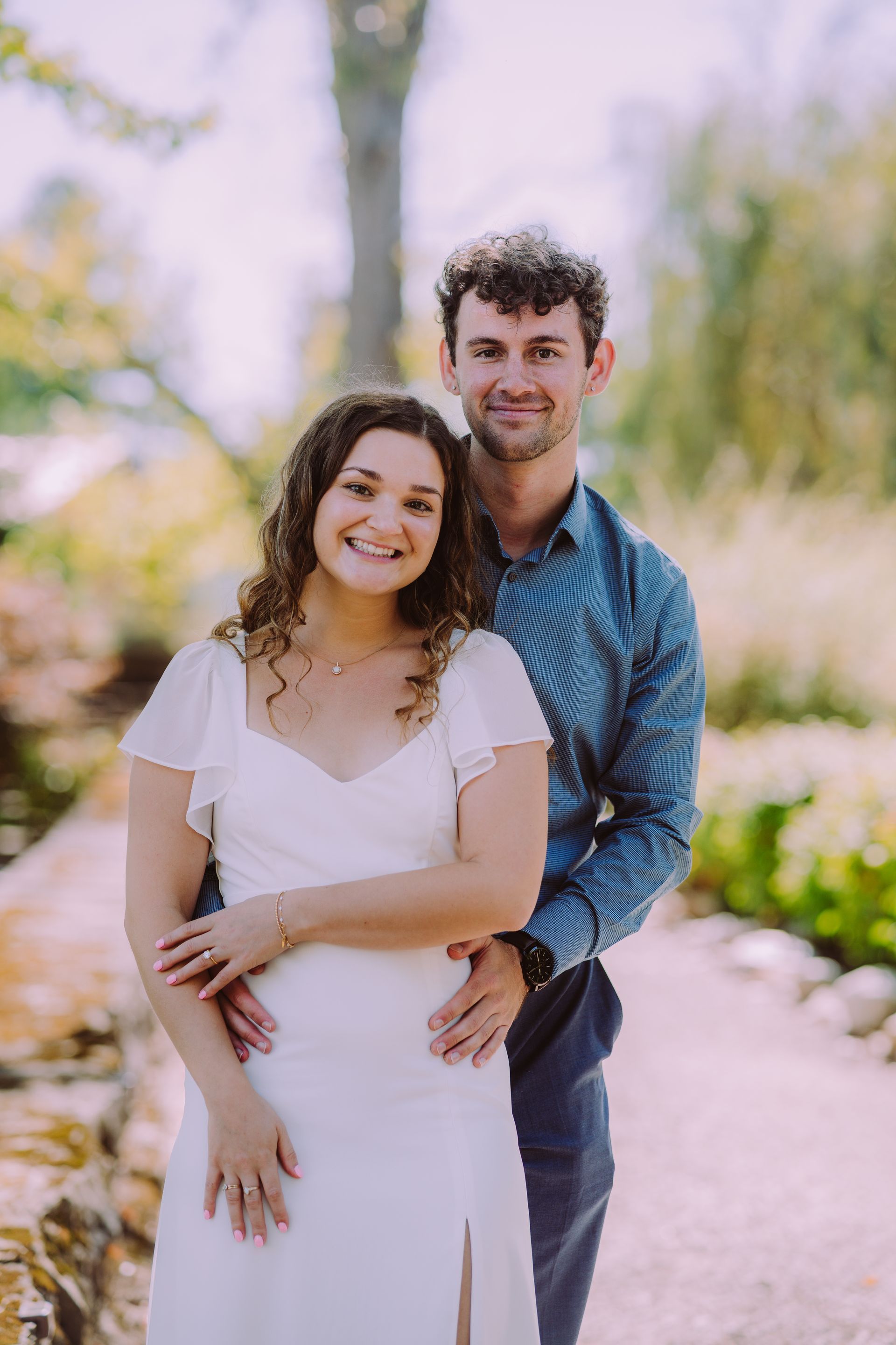 Couple smiles, poses on a path. Woman in white dress, man in blue shirt. Sunny outdoor setting.