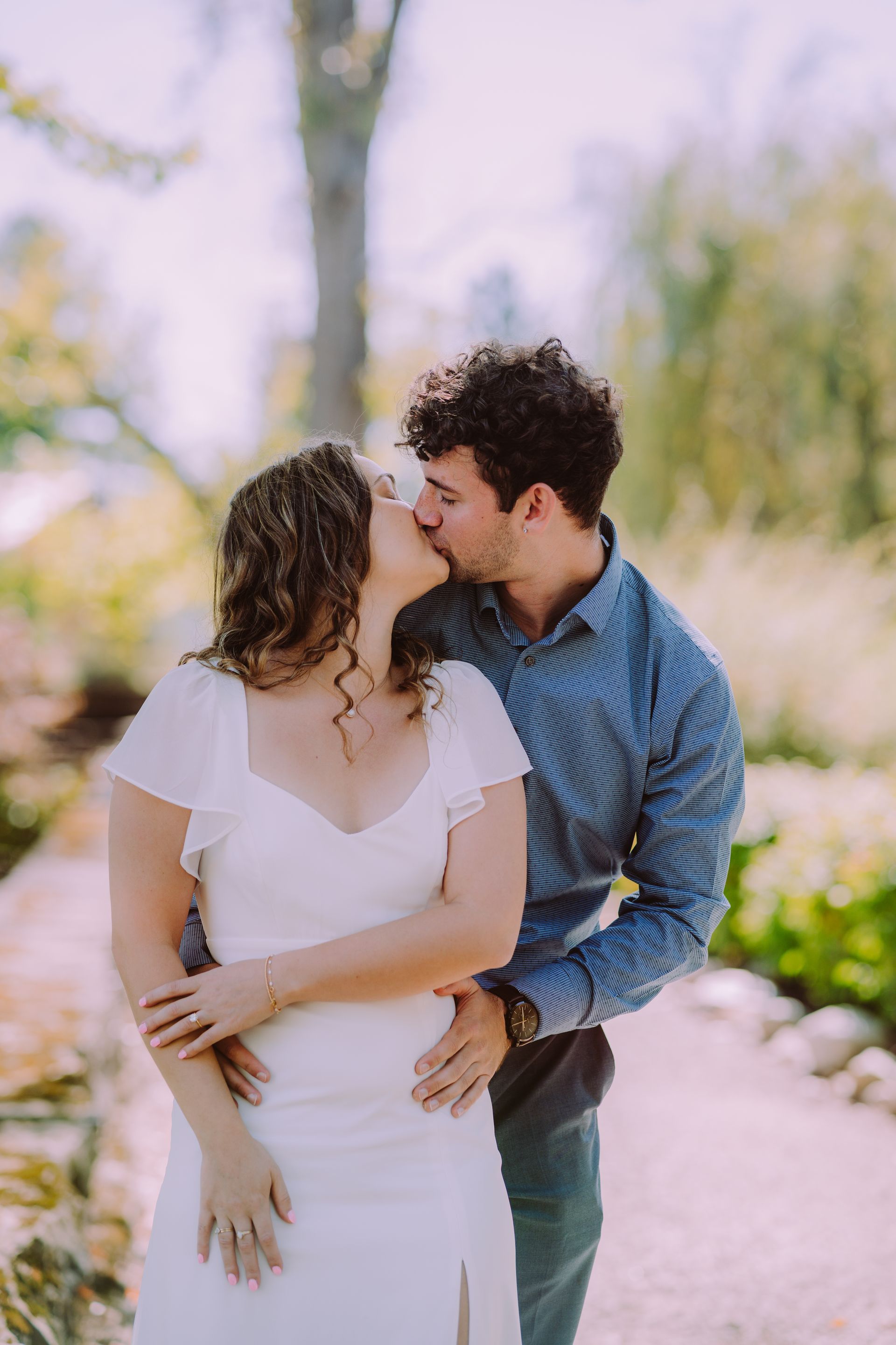 Couple kissing in a garden; woman in white dress, man in blue shirt, bright sunlight.