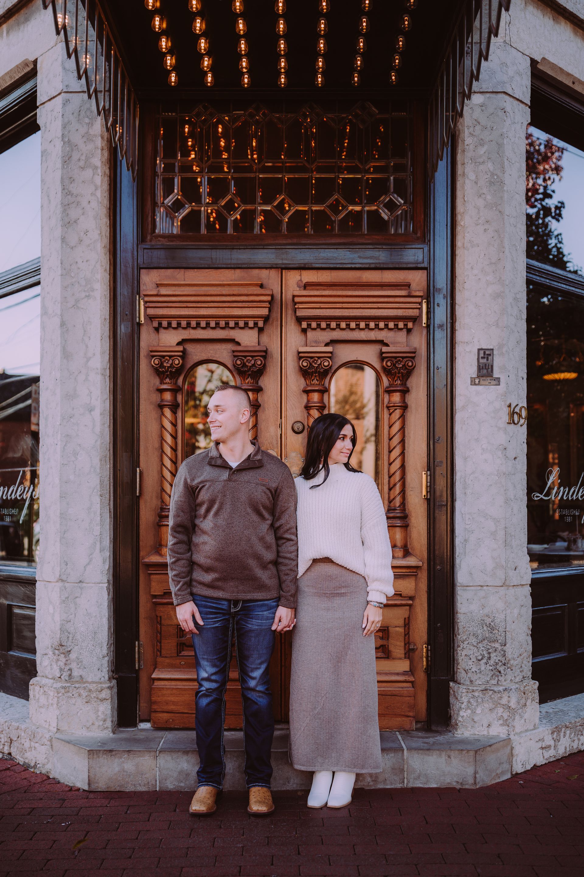 Couple holding hands in front of ornate wooden doors, stone pillars, and brick sidewalk.