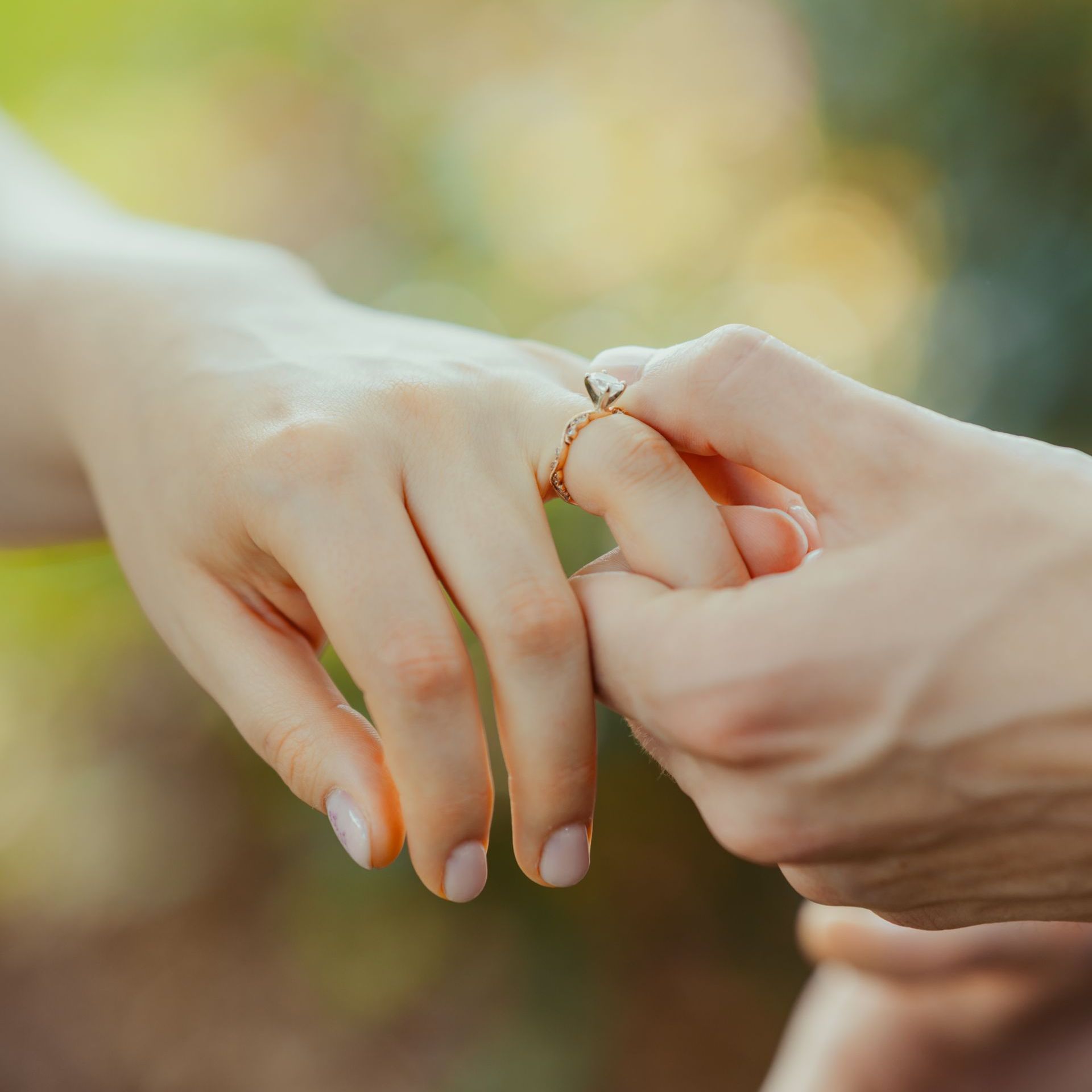 Person putting a gold ring with a diamond on another person's finger outdoors.
