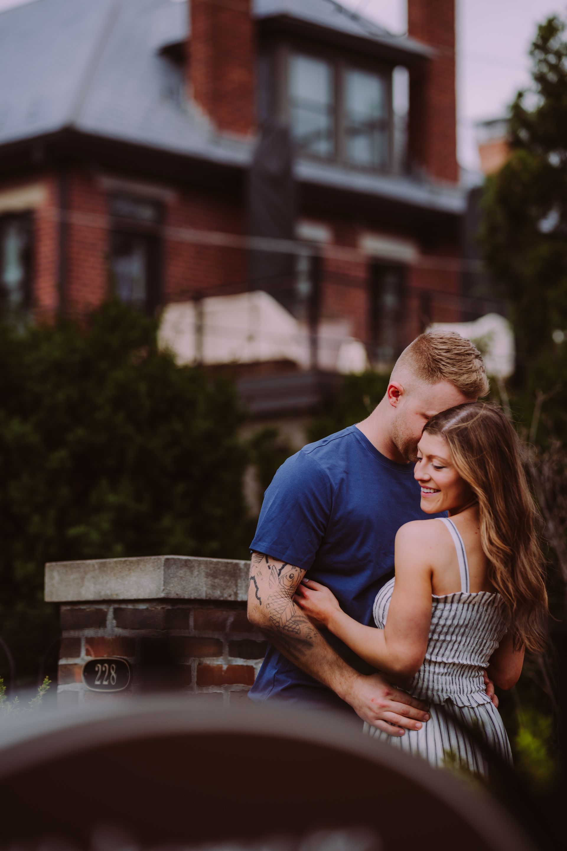 A couple embraces affectionately outdoors in front of a brick building with a stone pillar in the foreground.