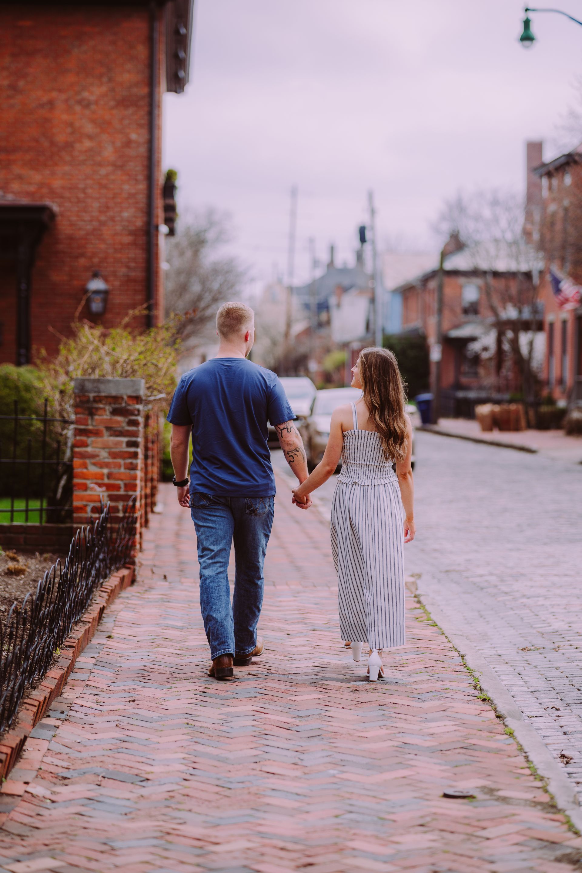 A couple holding hands walks away from the camera down a sidewalk lined with brick buildings and a cobblestone street.