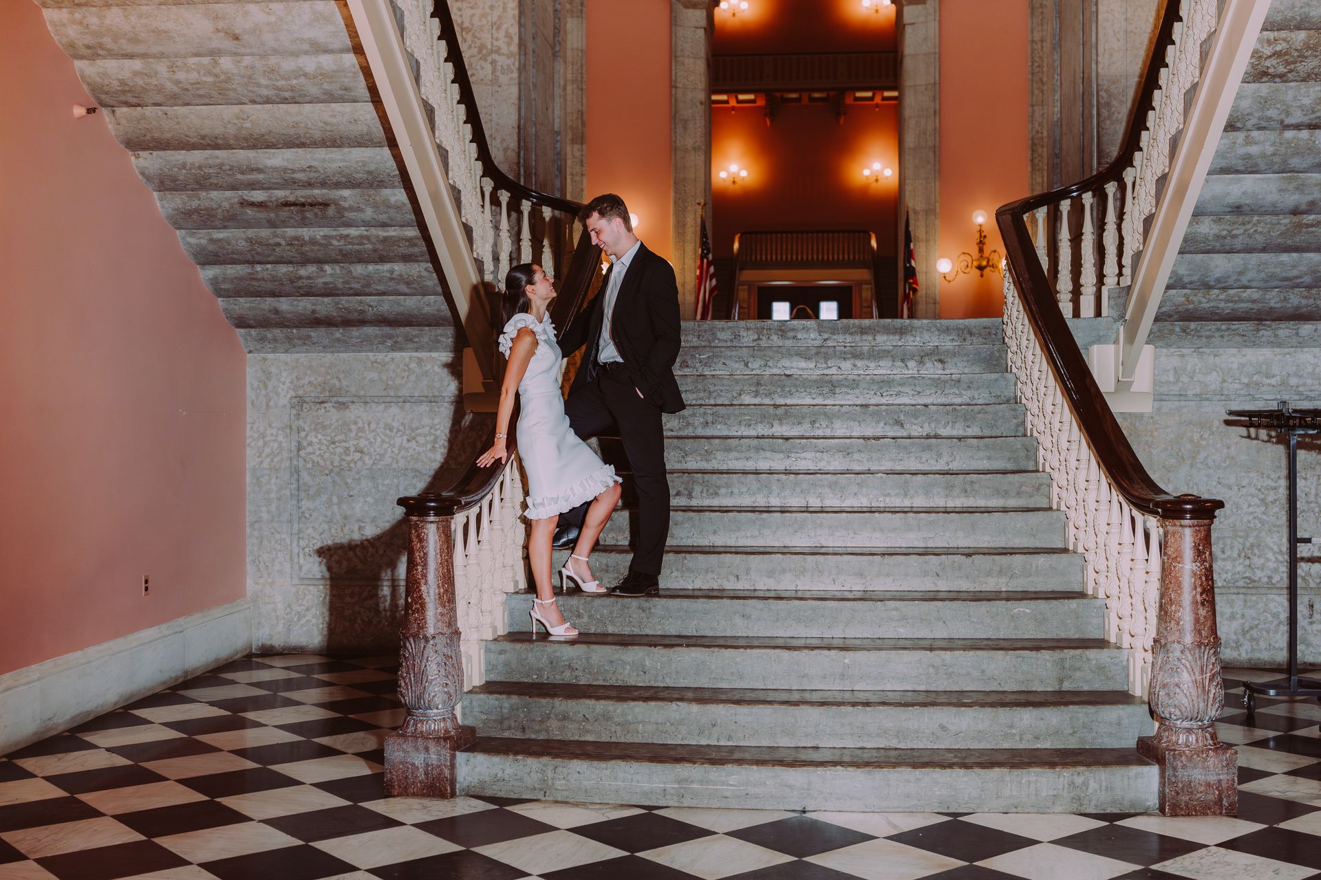 A couple standing on the stairs of a grand hall with marble floors and a black-and-white checkered floor pattern.