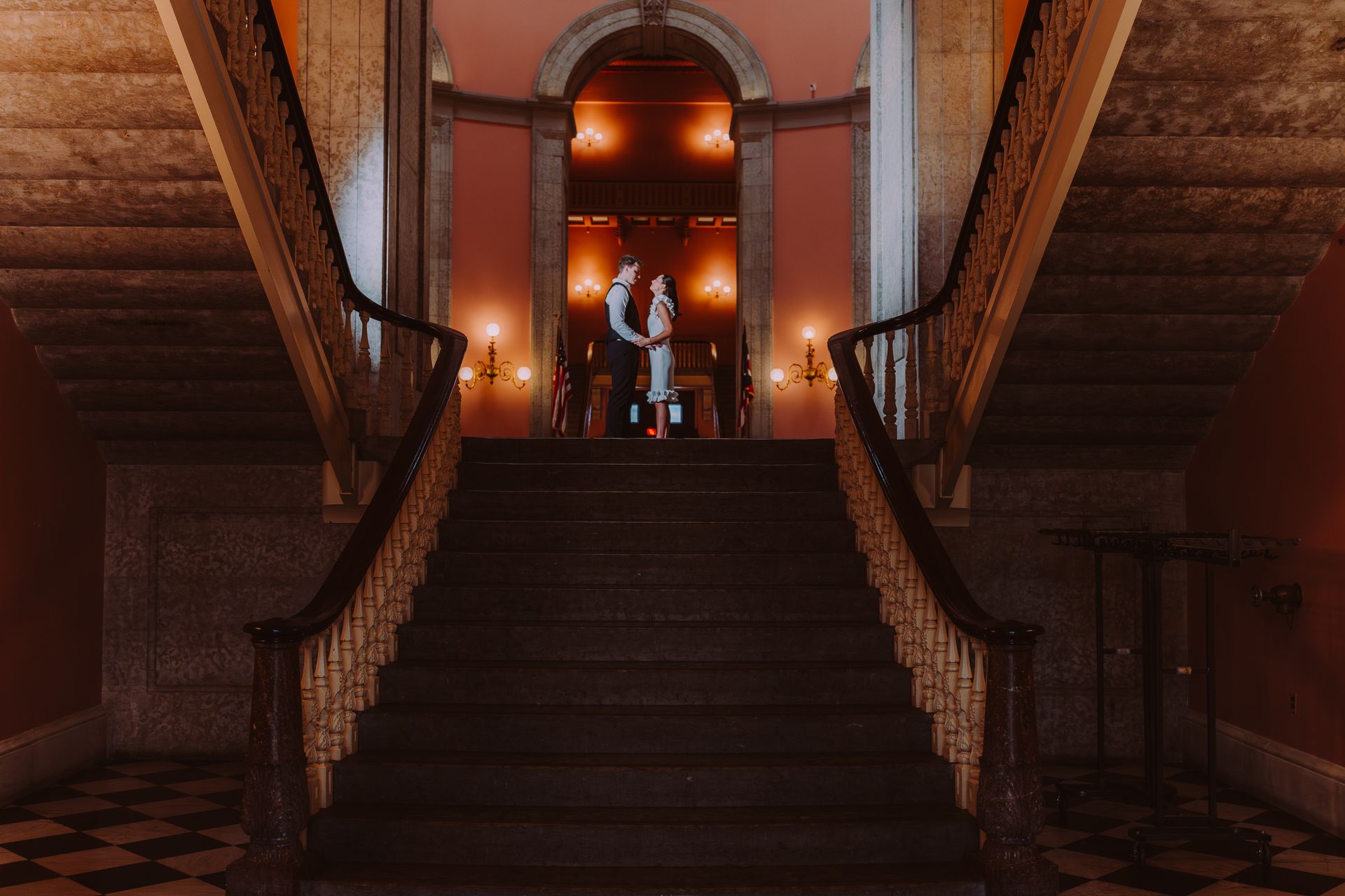 A couple stands together on a grand staircase inside a warmly lit, elegant building with tiled floors.
