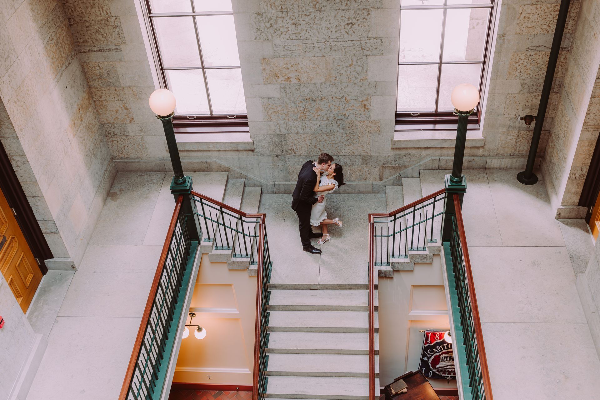 A couple embraces on a marble staircase in a grand hall with two tall windows and antique lamps.