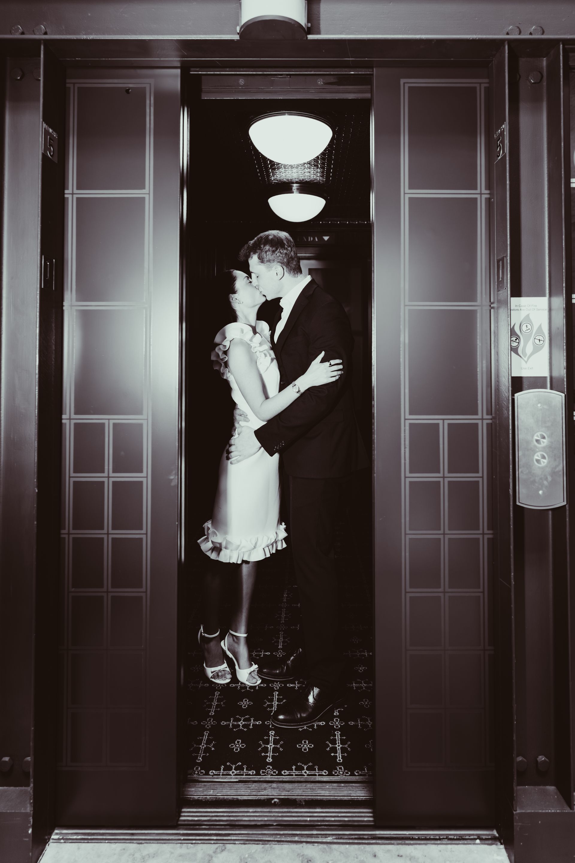 A couple in formal attire kissing inside a vintage elevator with patterned, dark metal doors and warm ceiling lights.