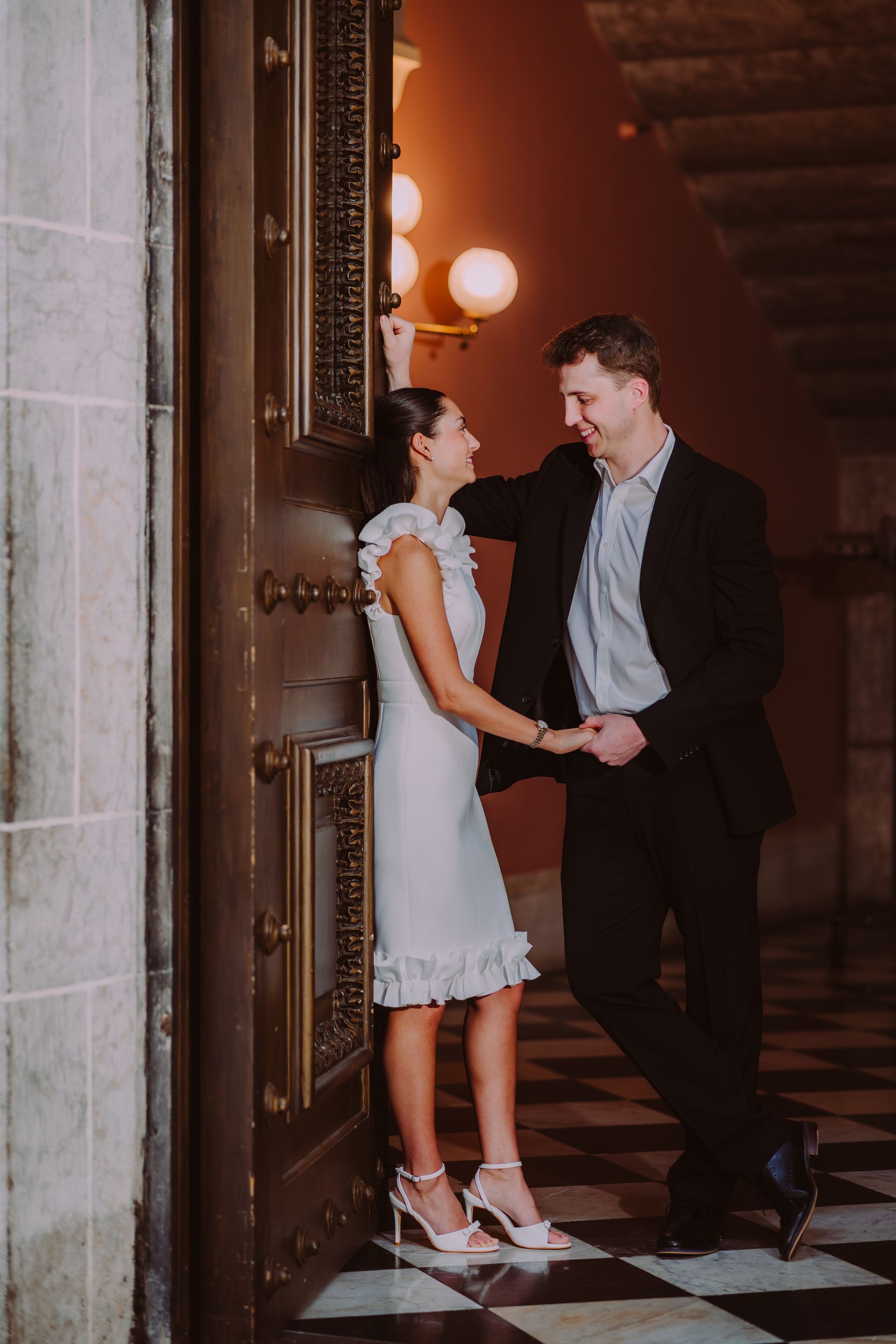 A couple in formal attire stands by a large wooden door in a hallway with a checkered floor, looking at each other.