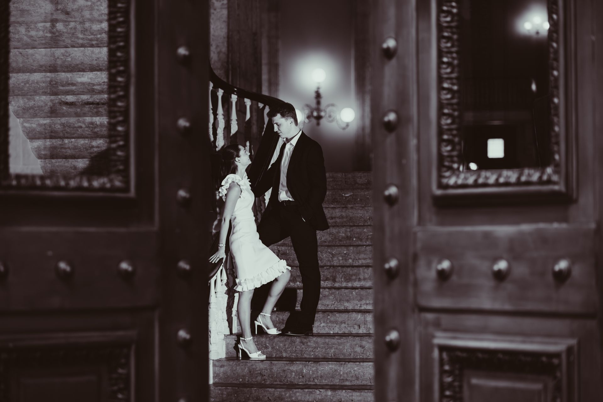 A couple stands on a grand staircase viewed through an open doorway in this black and white photograph.