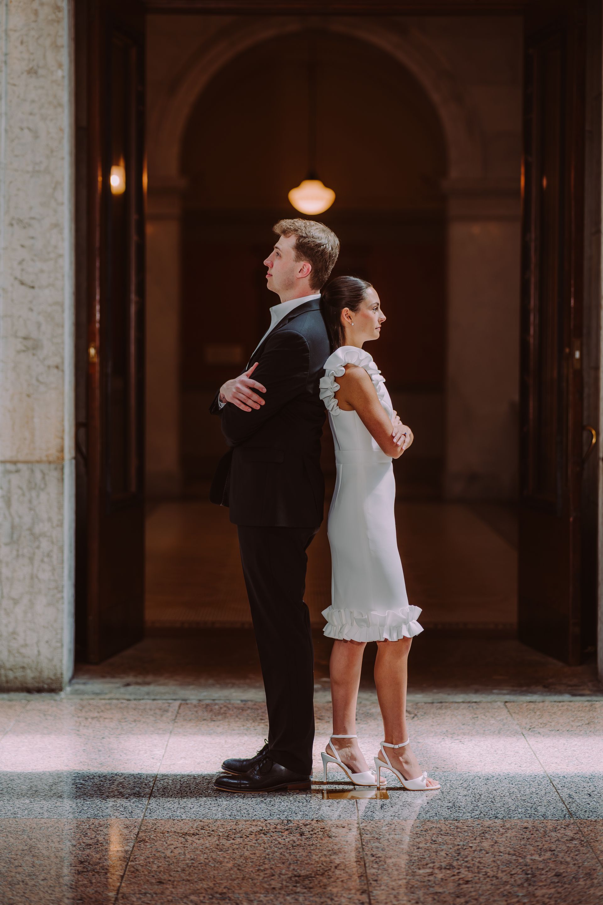 A couple stands back-to-back with arms crossed in an elegant, dimly lit marble corridor with an arched doorway.