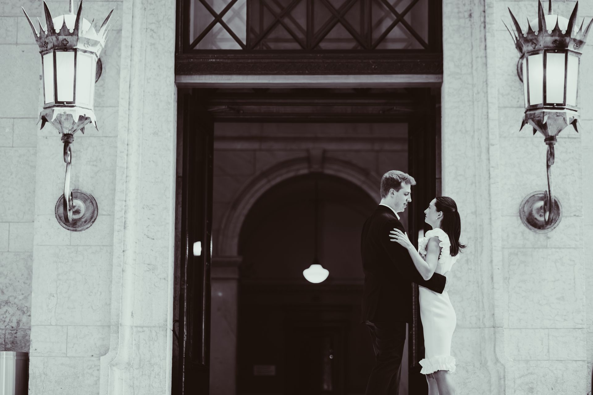 A couple dances outside a stone building entrance framed by two ornate vintage lanterns in a black-and-white photograph.