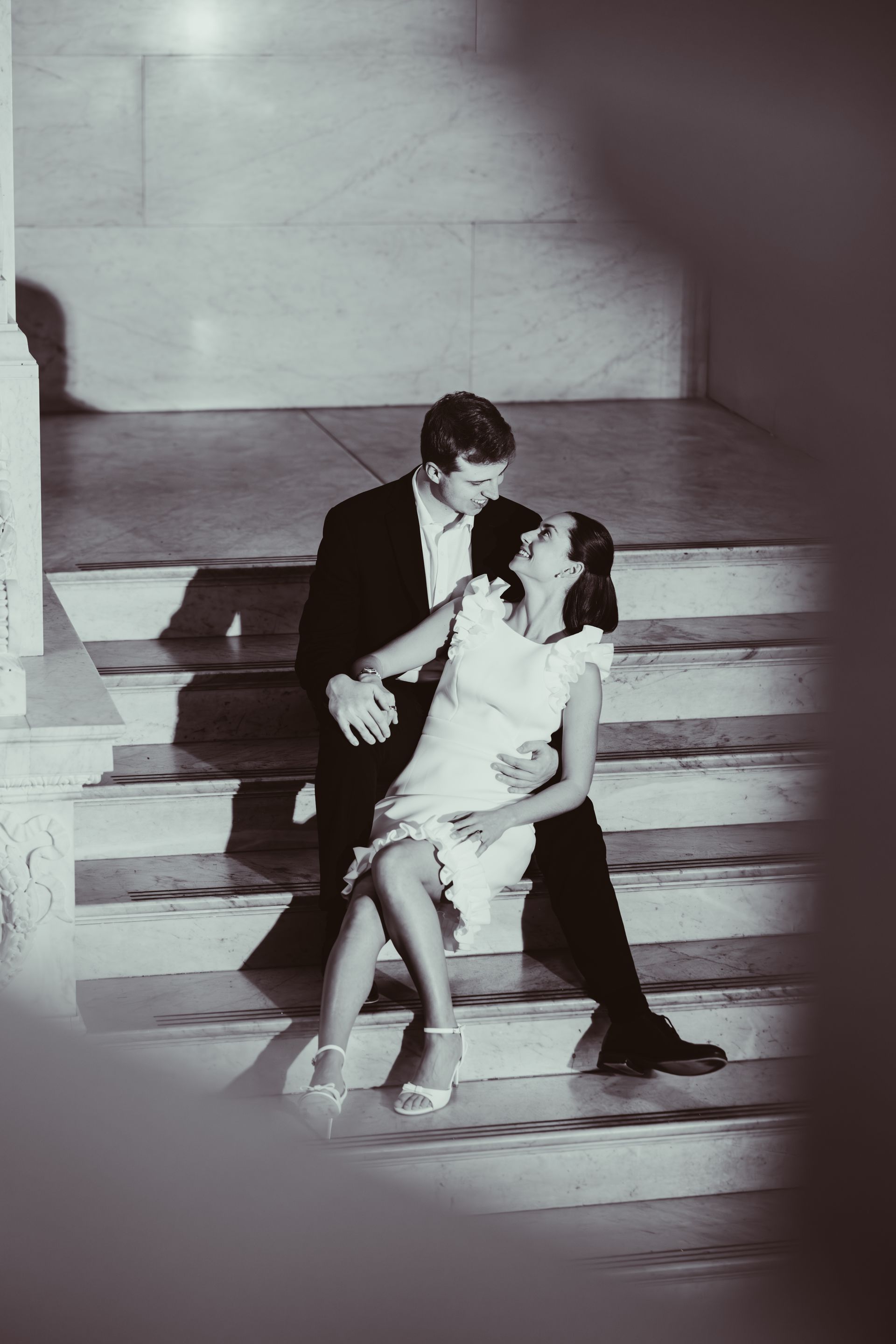 A couple sits on marble stairs, the man in a dark suit embracing the woman, who wears a light-colored dress.