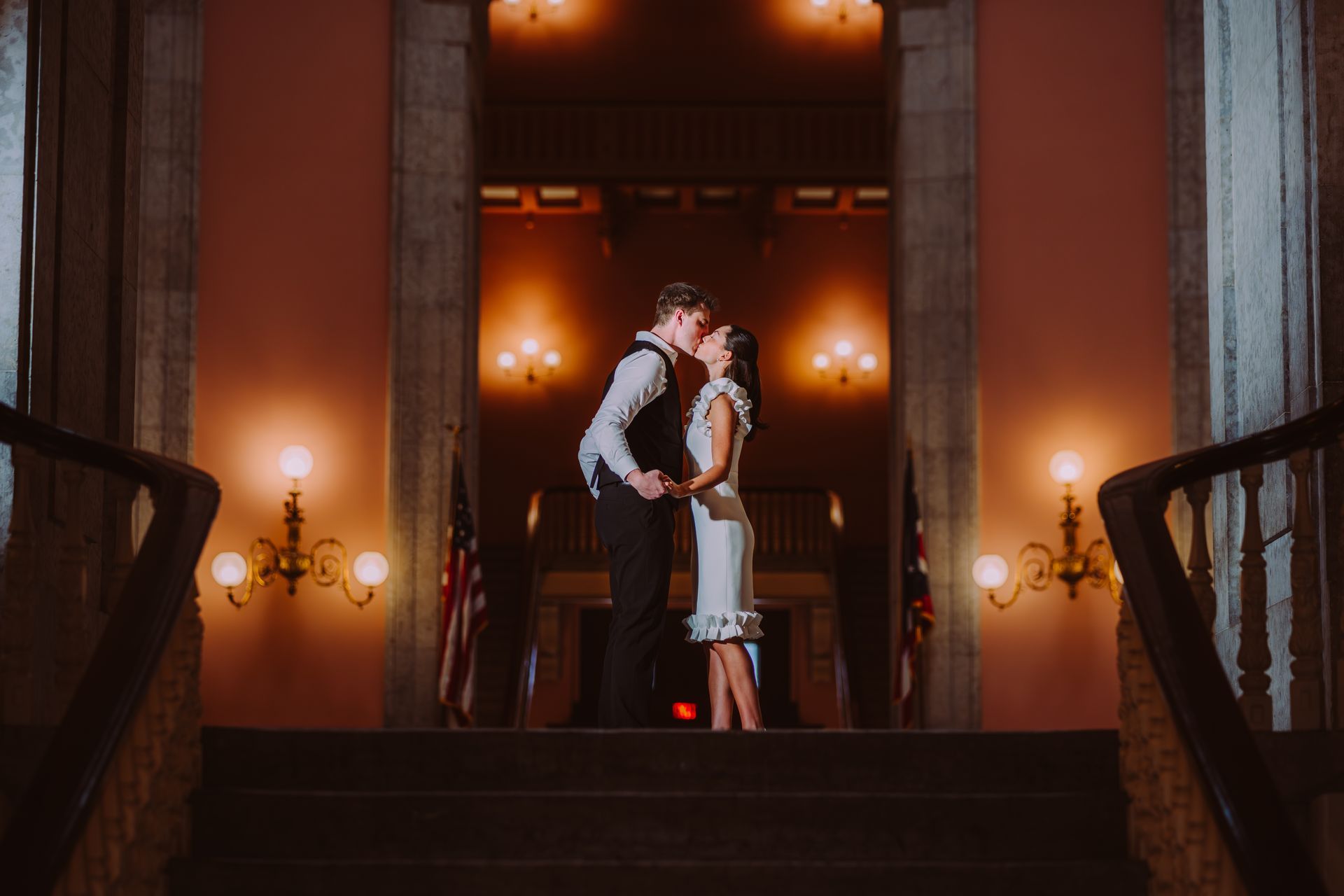 A couple kissing while standing on a staircase in a grand, dimly lit hallway with warm wall sconces.