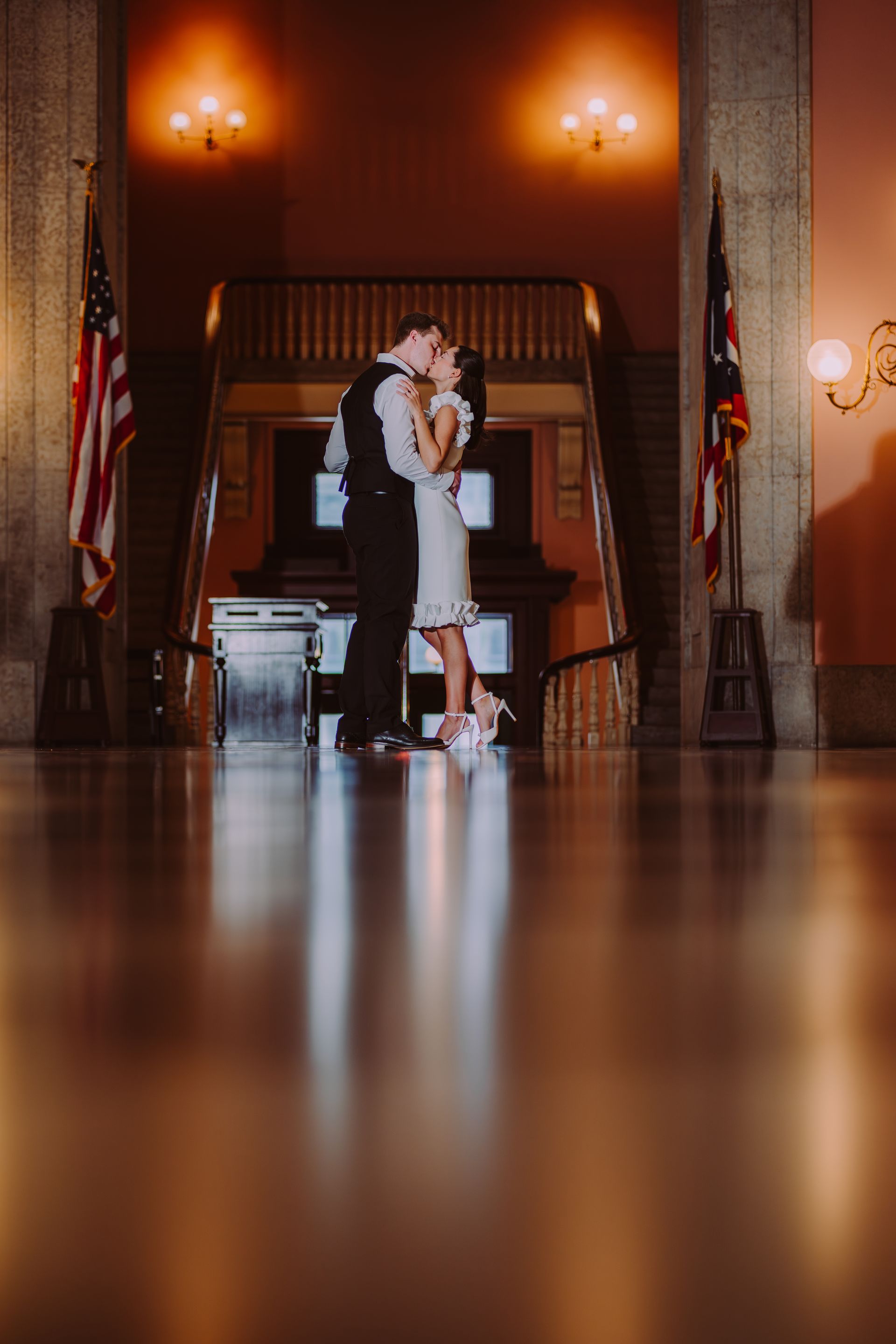 A couple in formal attire embracing and kissing in a grand hall, framed by American flags and warm ambient lighting.