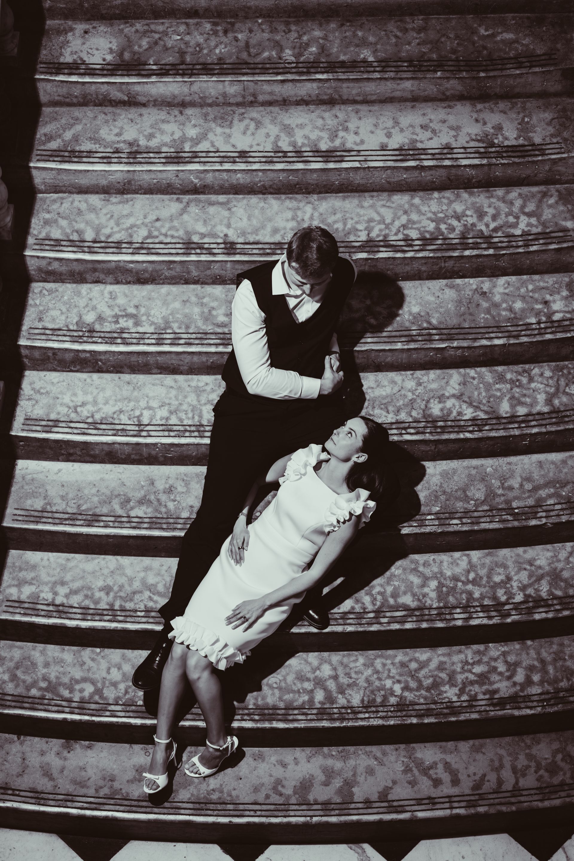 A couple in formal attire sitting on grand stairs, with one person leaning back toward the other in a romantic pose.
