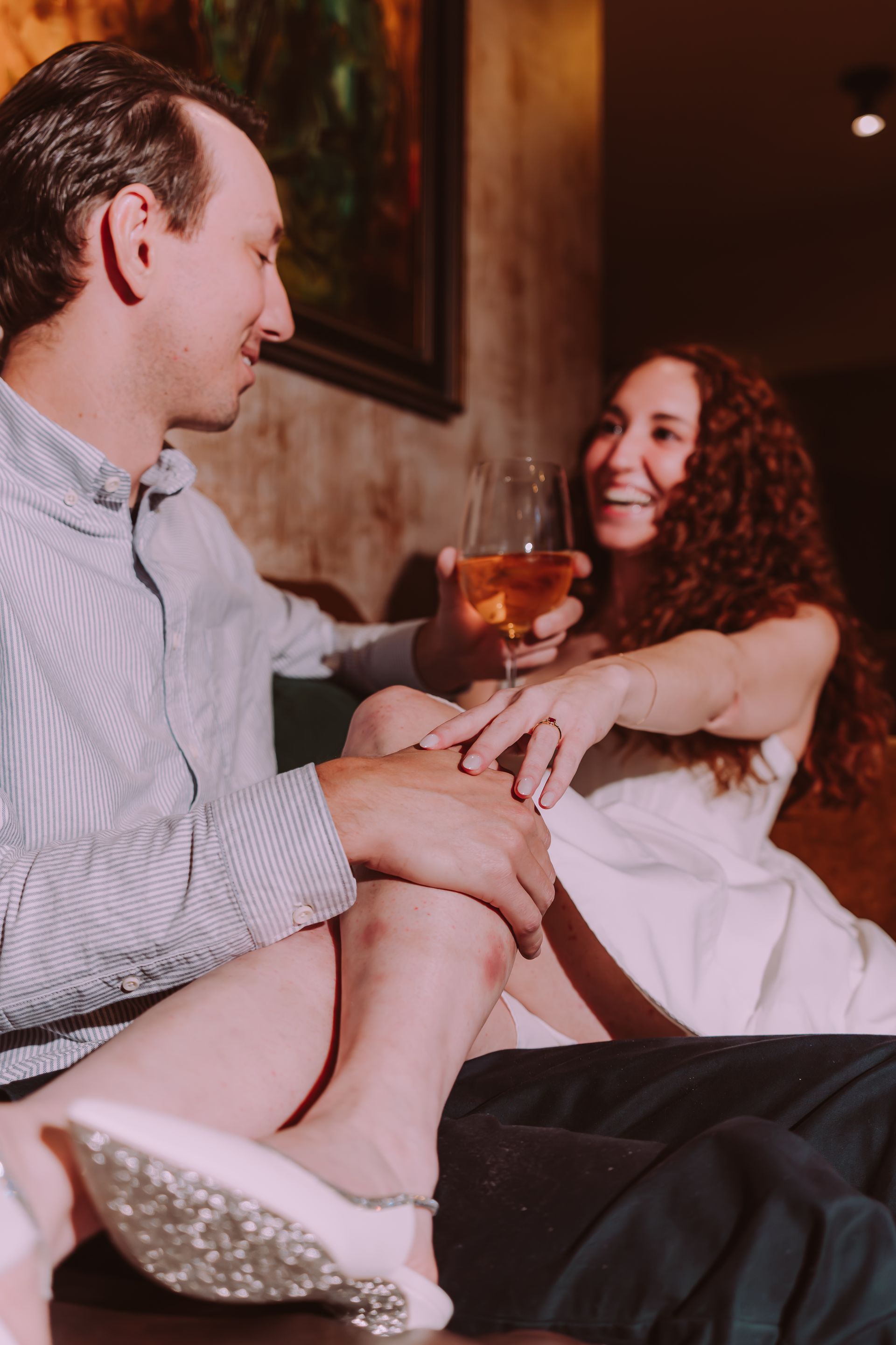 Man and woman smiling, sitting close. Woman holds wine, wears white dress. Romantic setting.