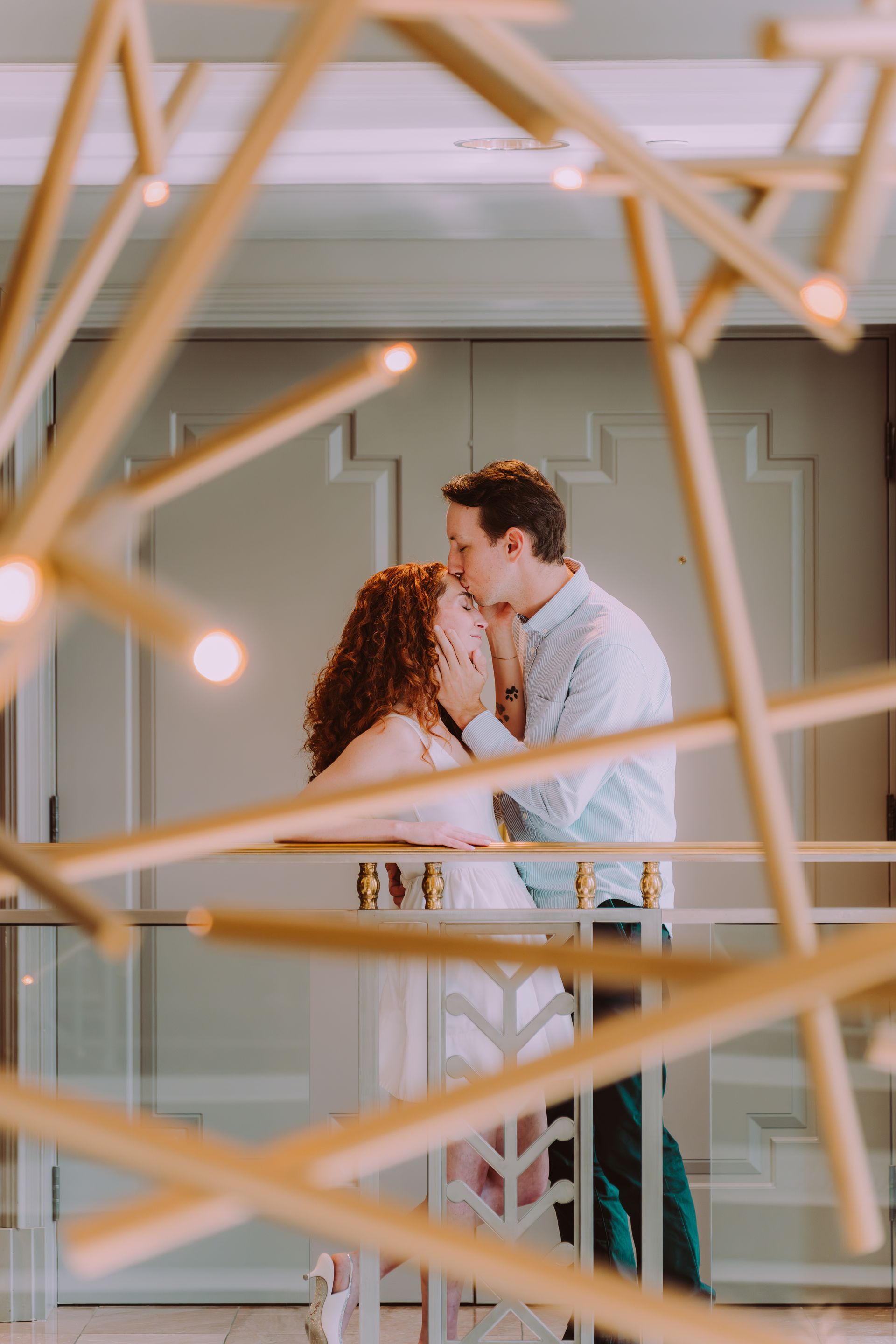 Couple embracing on a balcony. Man kisses woman's forehead. Gold geometric chandelier in foreground.