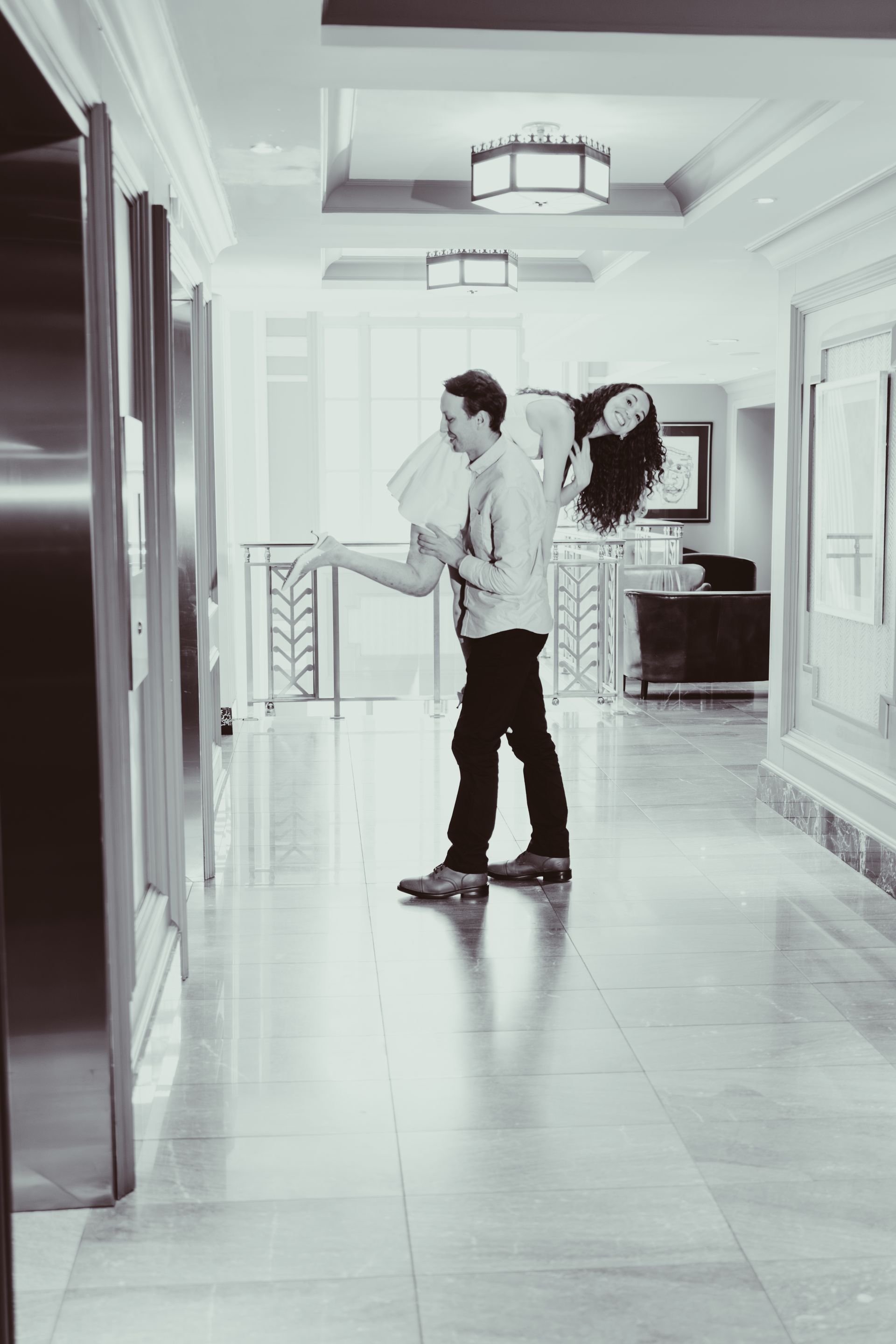 Man carrying woman in a hallway, pressing elevator button. Black and white.