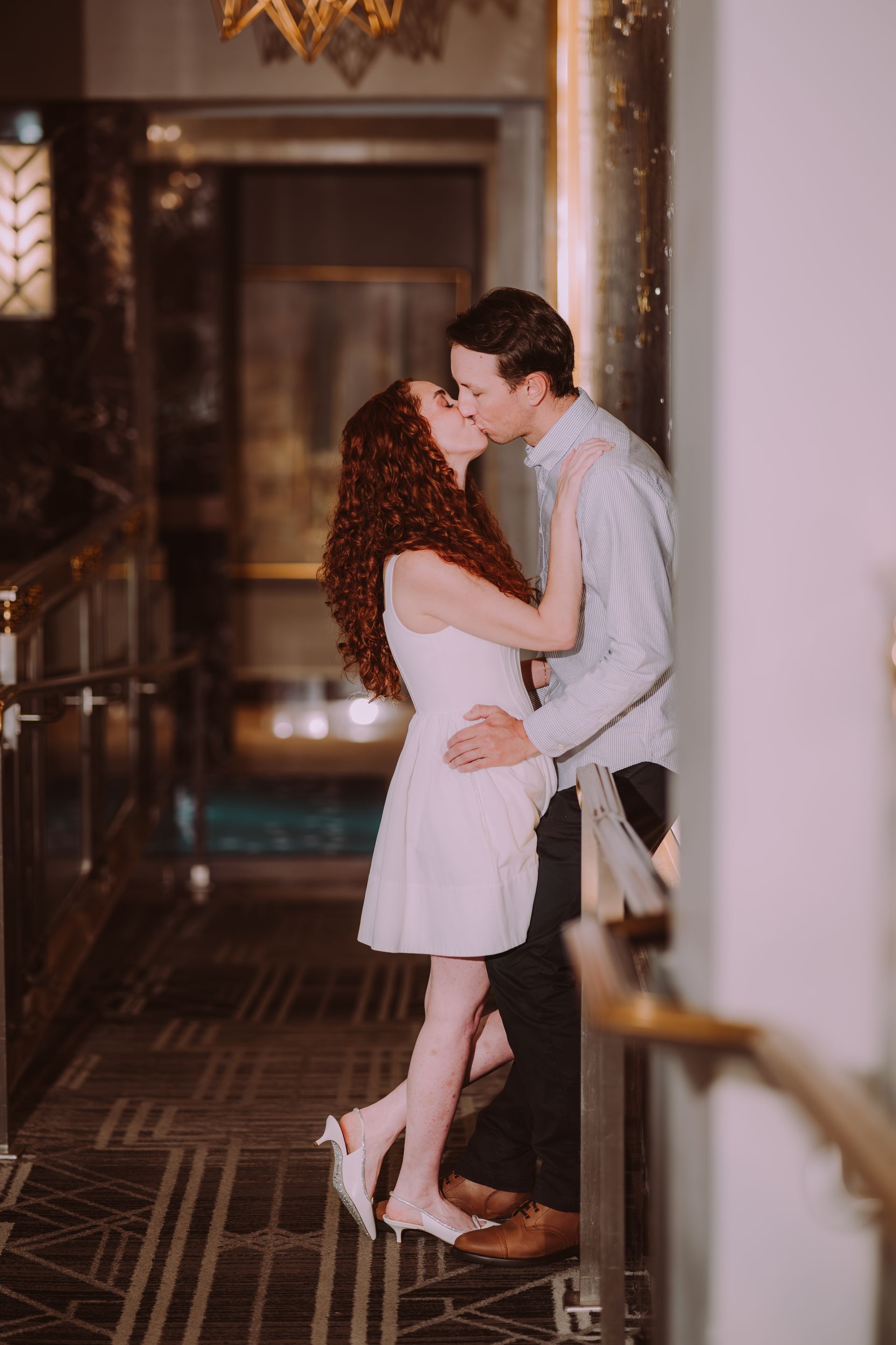 Couple kissing in a hallway. Woman in white dress, red hair; man in blue sweater. Dim lighting, golden accents.