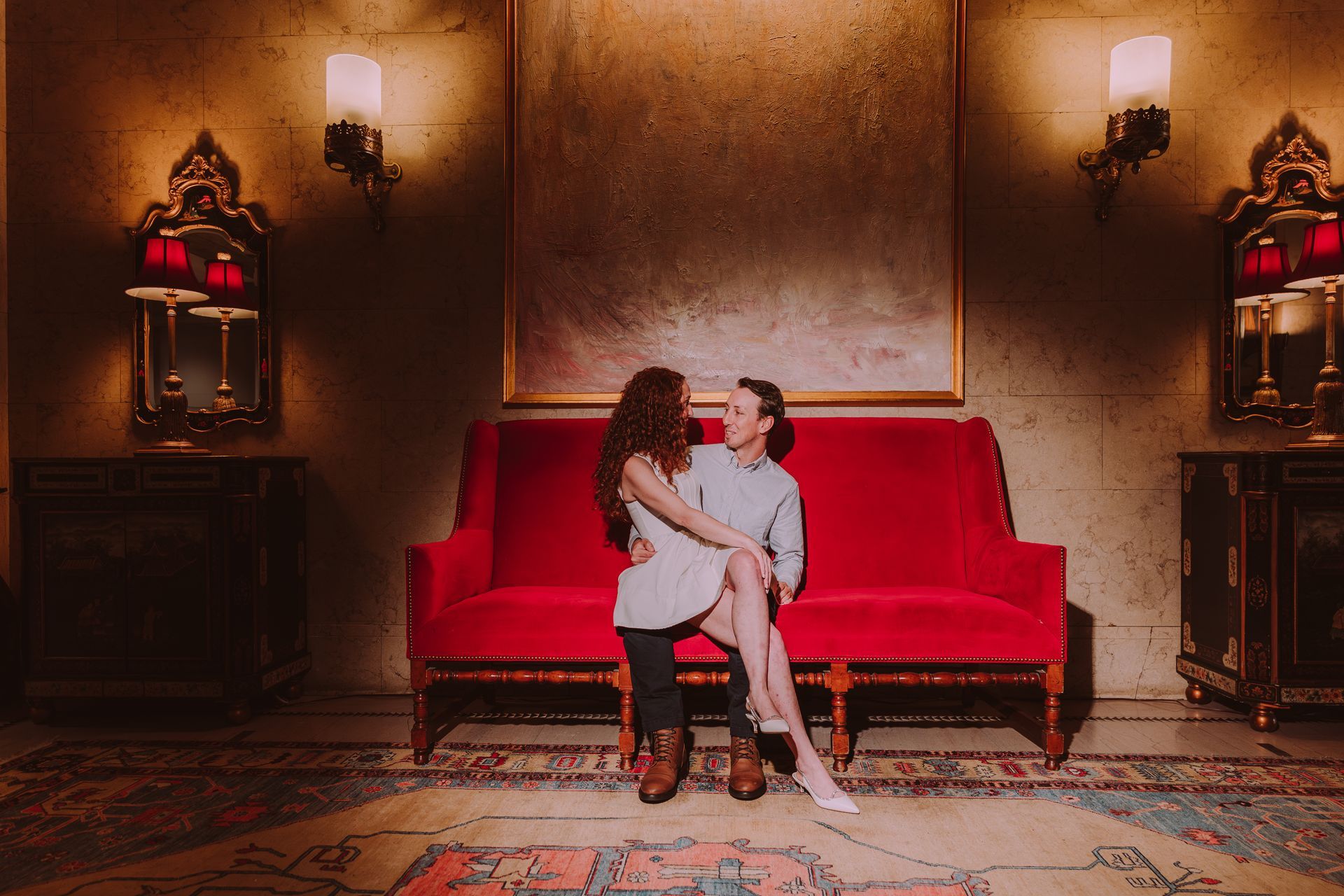Two women embrace on a red couch in a room with ornate decor; one has red hair.