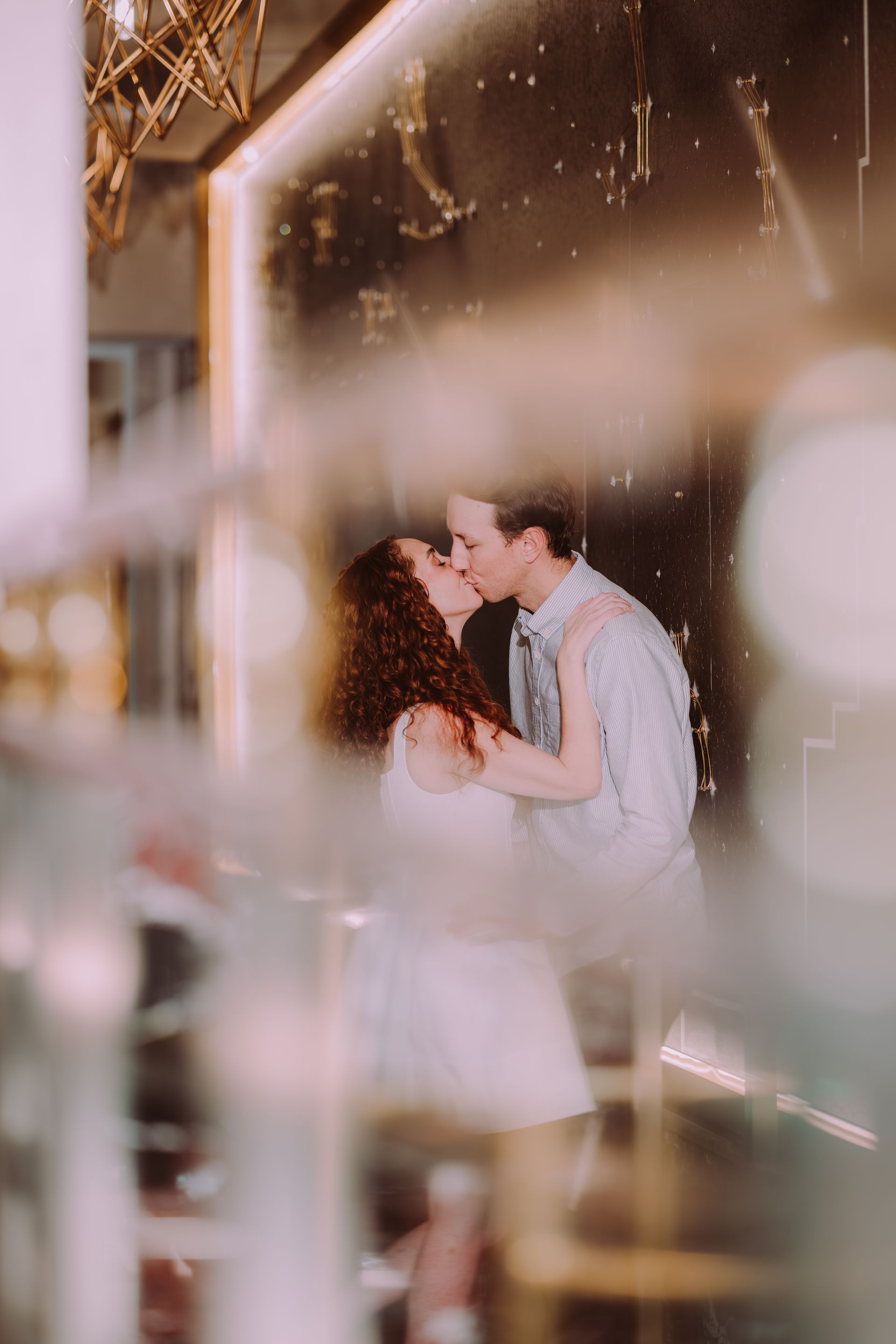 Couple kissing, embraced, on a staircase. She has red curly hair, he wears a blue shirt. Blurred foreground.