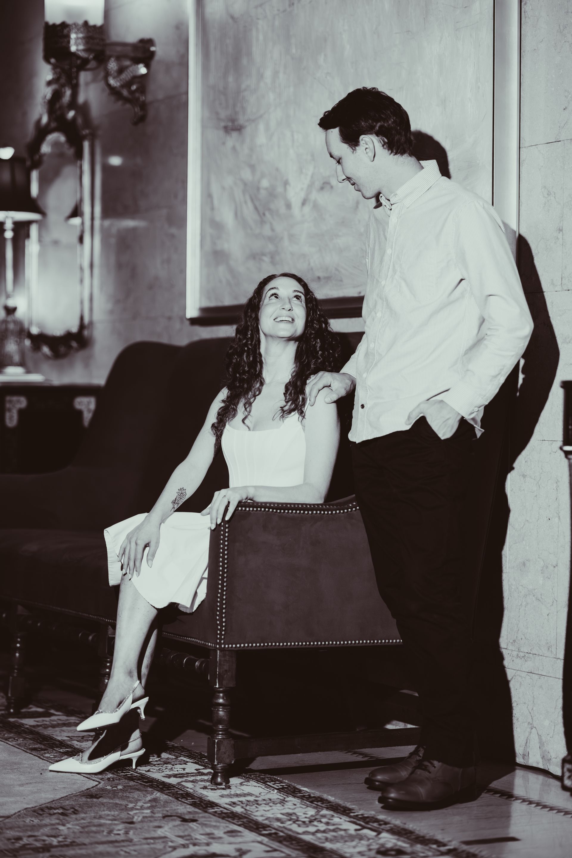 Man and woman in vintage room. She sits, smiling up at him. He stands, hand on her shoulder.