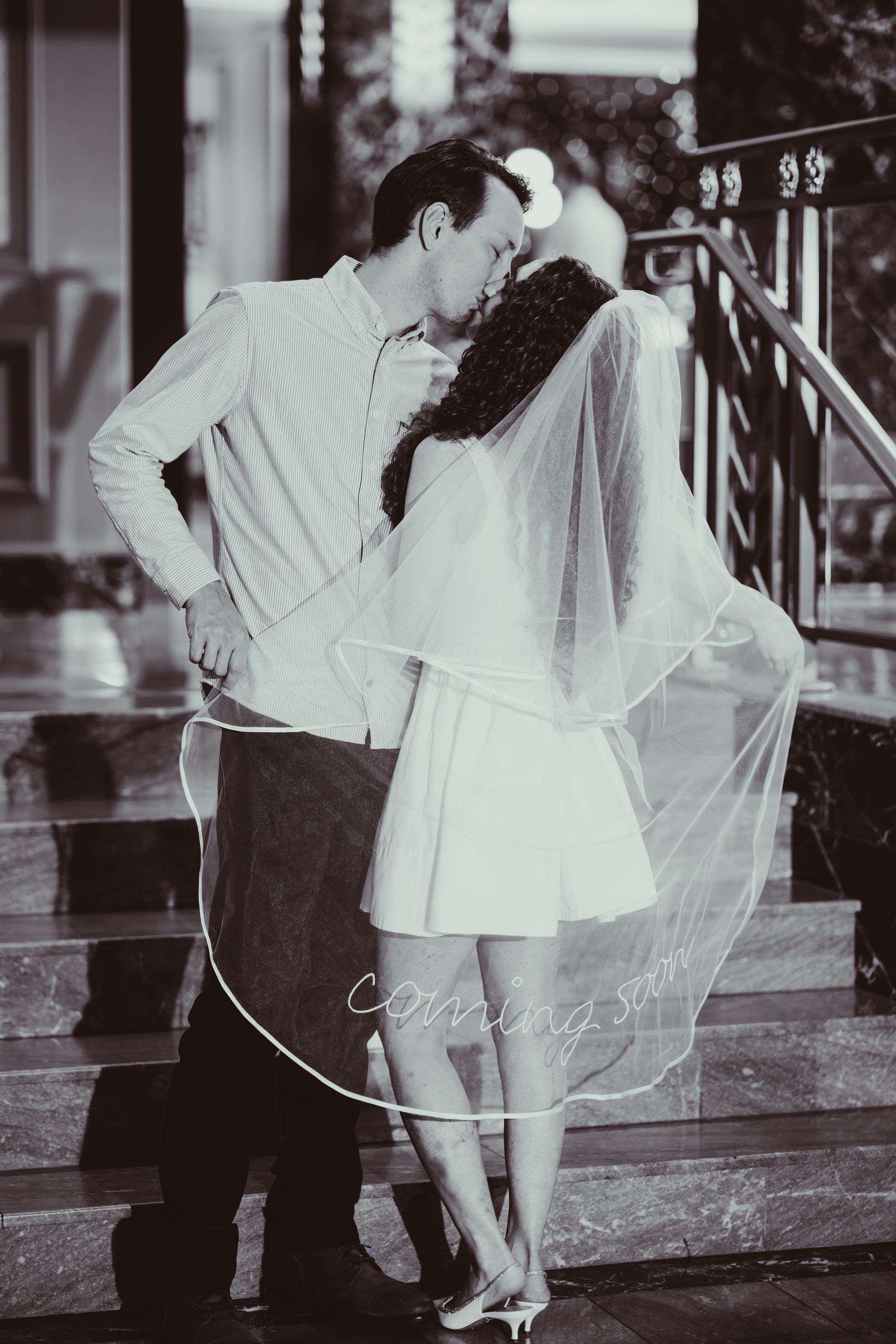 Newlyweds kissing on steps; bride in short dress and veil, groom in shirt. Black and white.