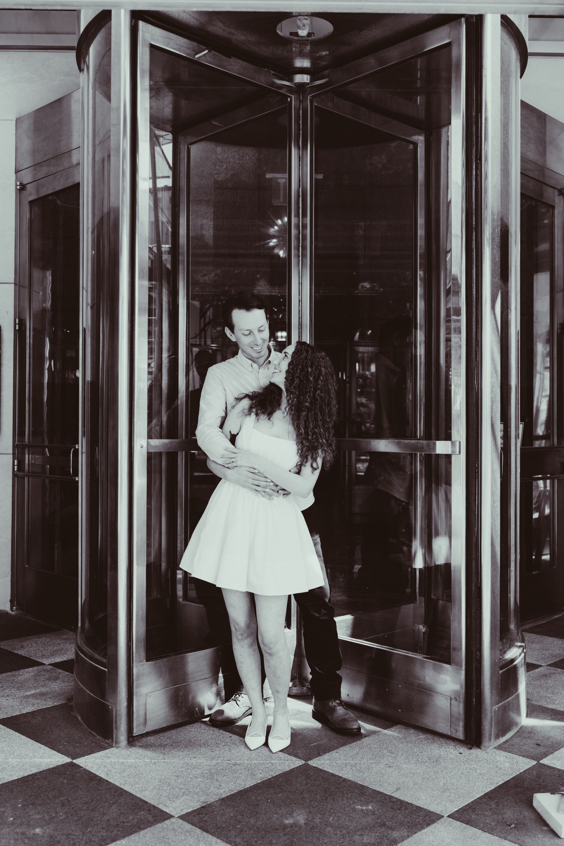 A couple embraces in a revolving door. The woman wears a dress and heels; the man, a dark suit. Black and white.