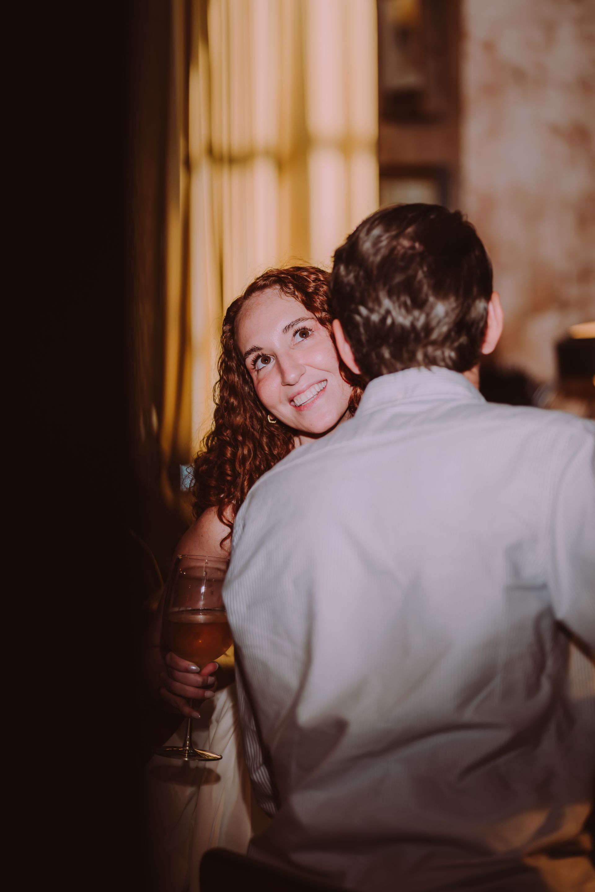 Woman with red curly hair smiles at the man in front of her, holding a drink. Indoors, warm lighting.