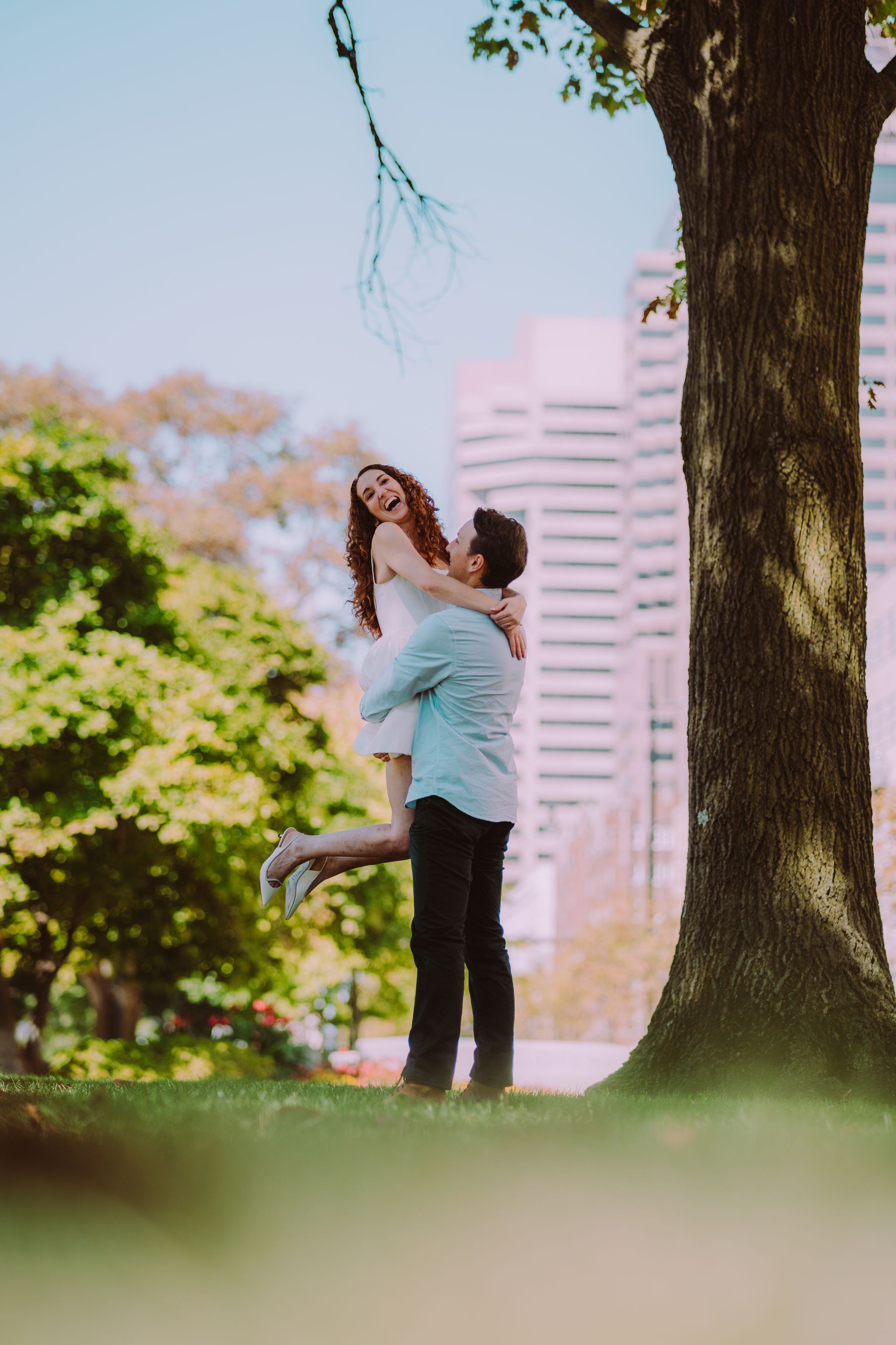 Man lifting woman, both smiling, under a tree in a park, buildings in background.