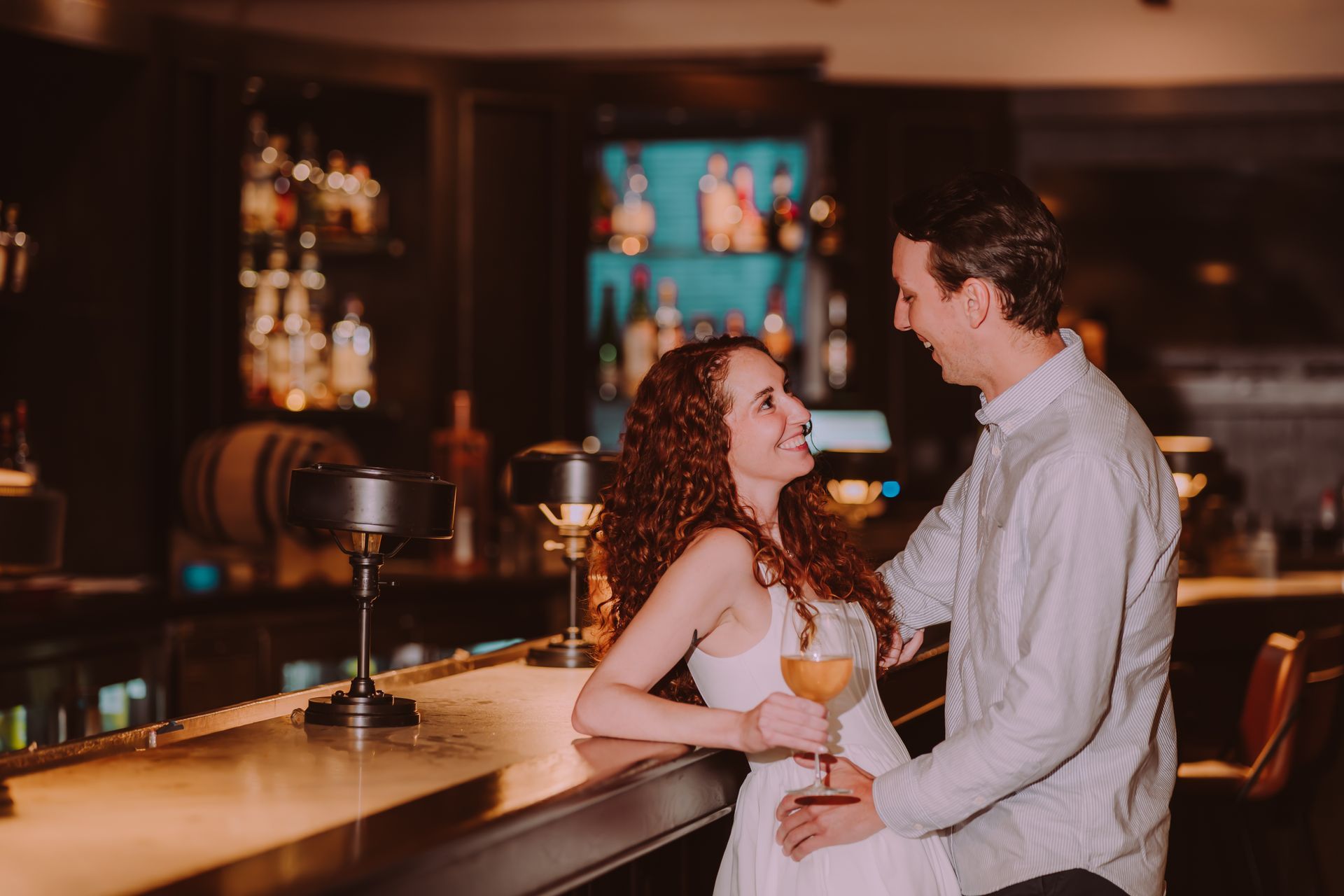 Couple at a bar, smiling and gazing at each other. Woman in white dress, man in a button-down.